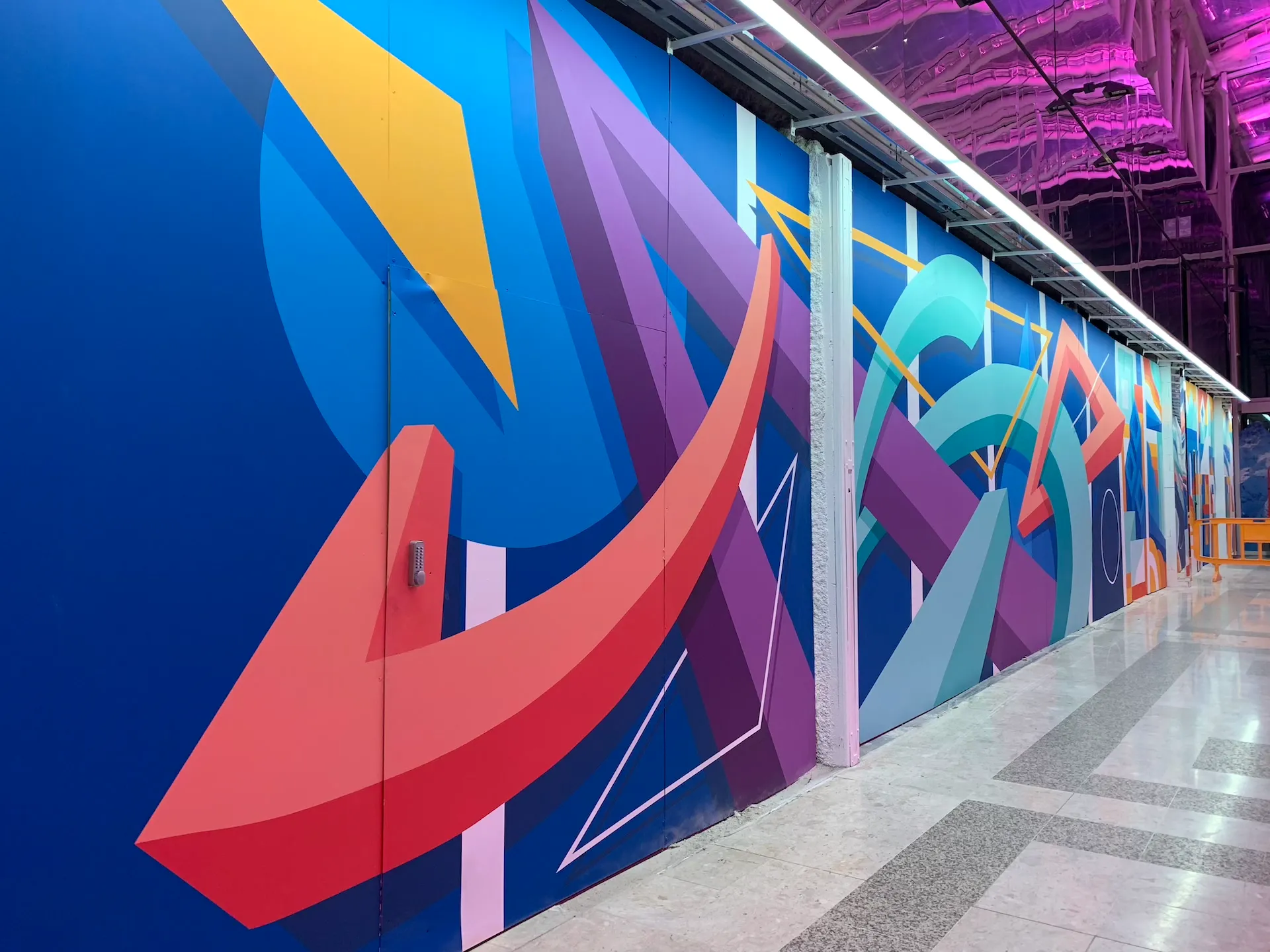 Couloir décoré de fresques géométriques multicolores sur fond bleu, sous un éclairage violet.