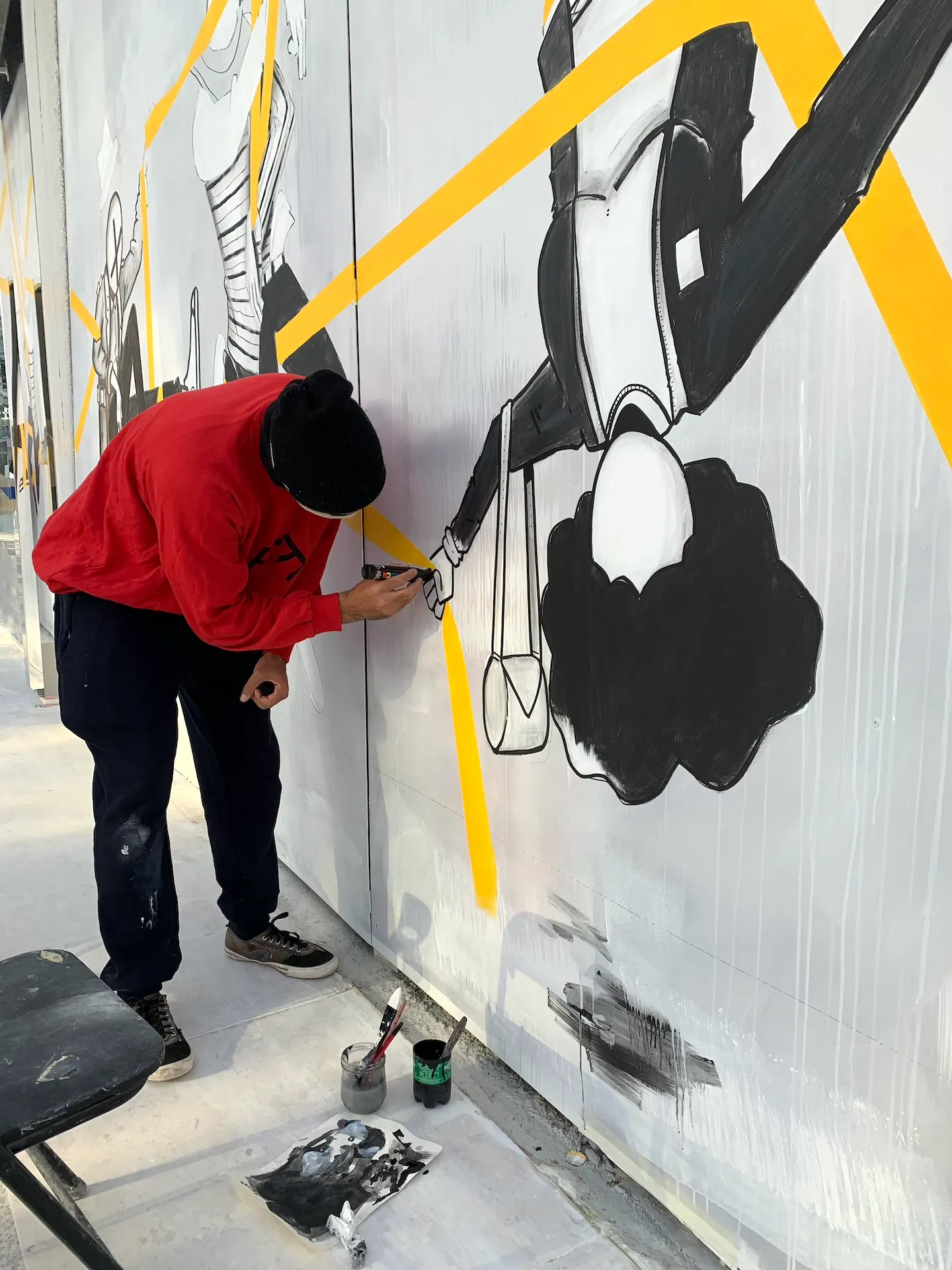 Artiste en train de peindre une fresque en noir et blanc avec des formes graphiques sur fond de bandes jaunes.