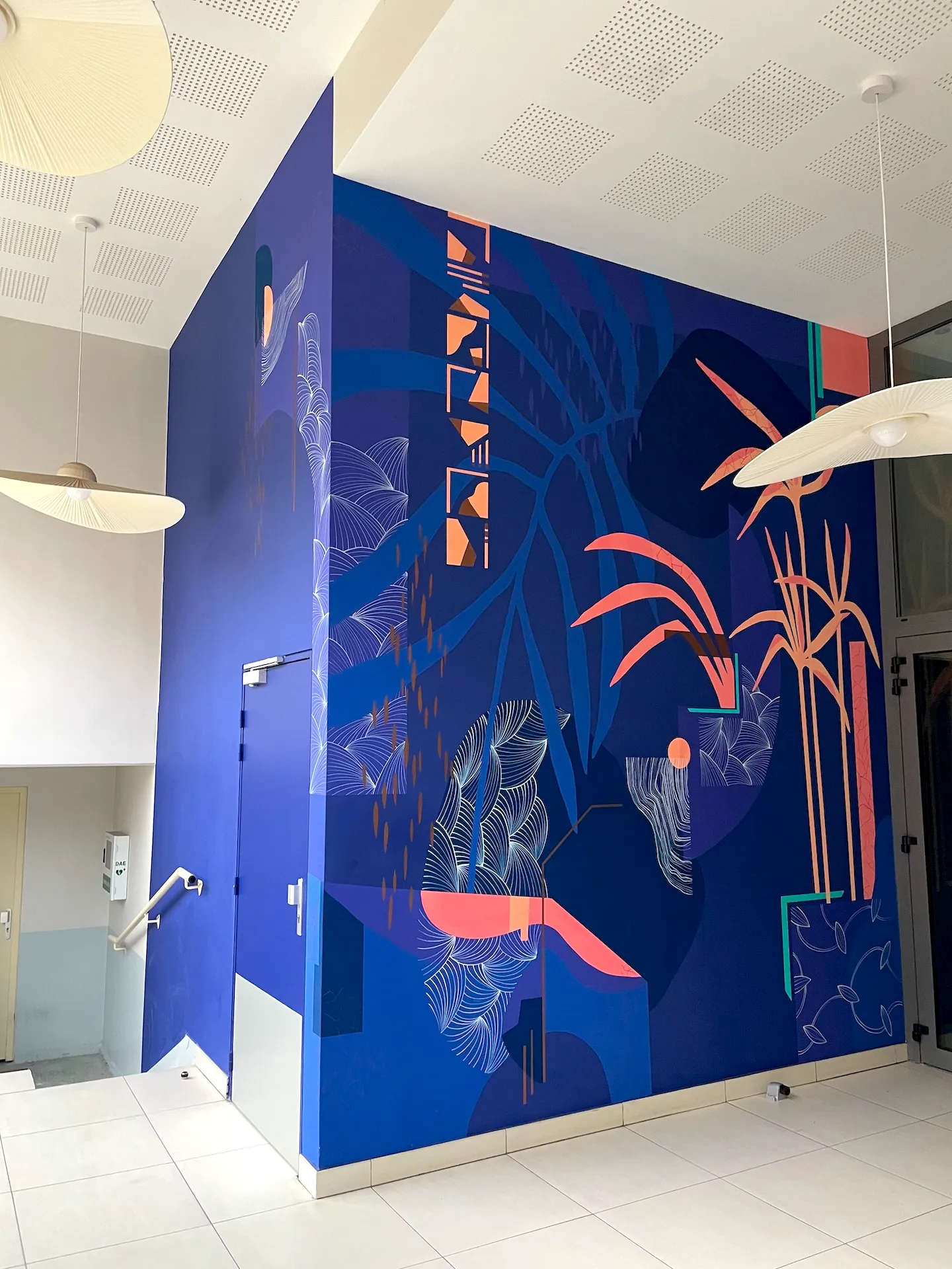 Vue d'une fresque habillant un angle de mur dans un hall, totalement bleue et ponctuée d'éléments graphiques et végétaux roses et blancs.