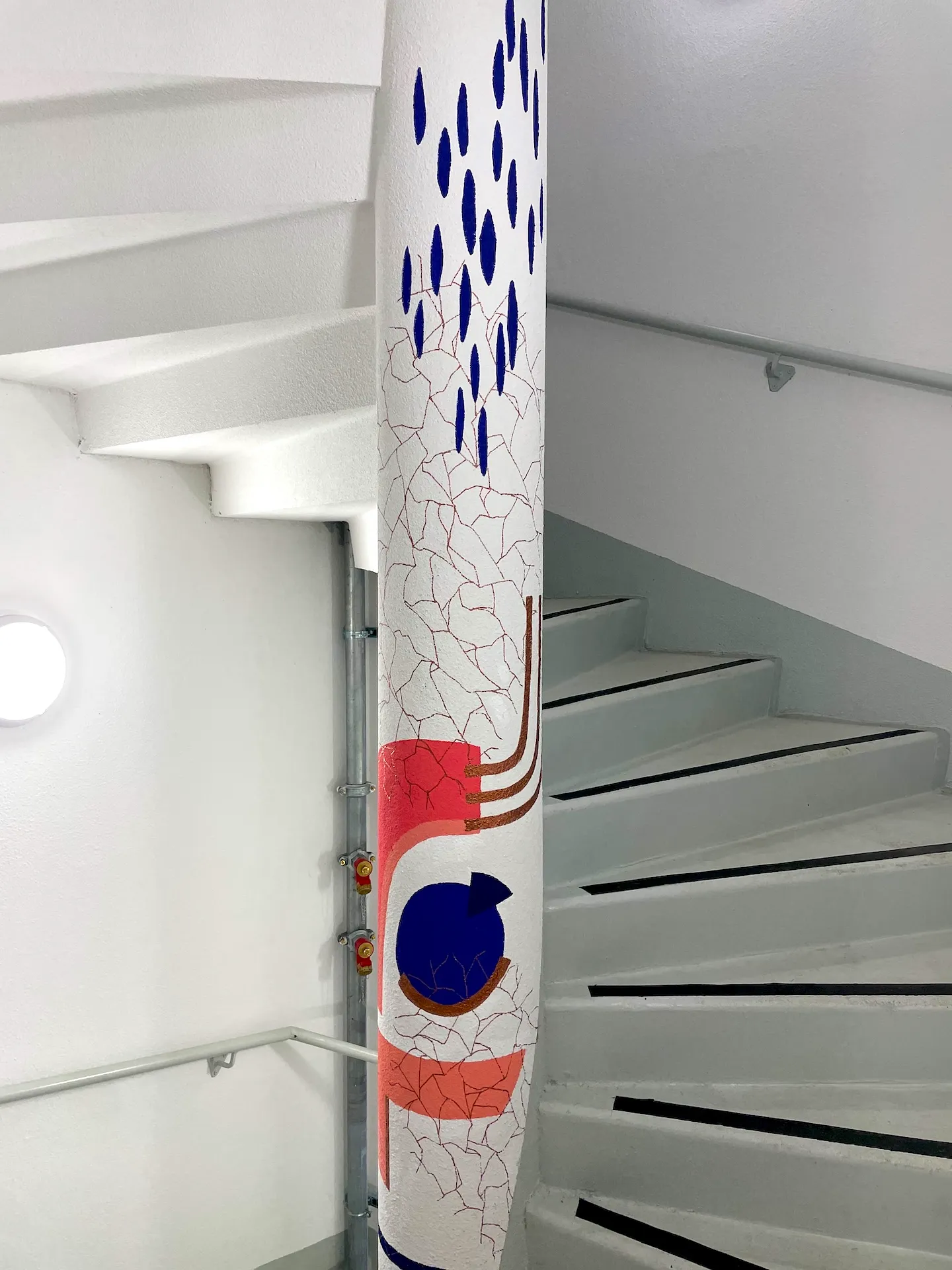 Éléments graphiques et végétaux bleus et roses sur un pylône dans une cage d'escaliers blanche.