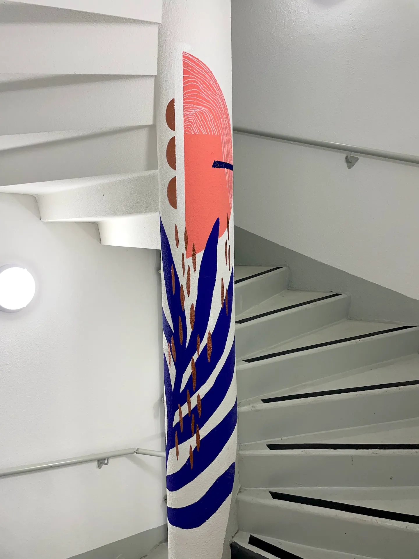 Éléments graphiques et végétaux bleus et roses sur un pylône dans une cage d'escaliers blanche.