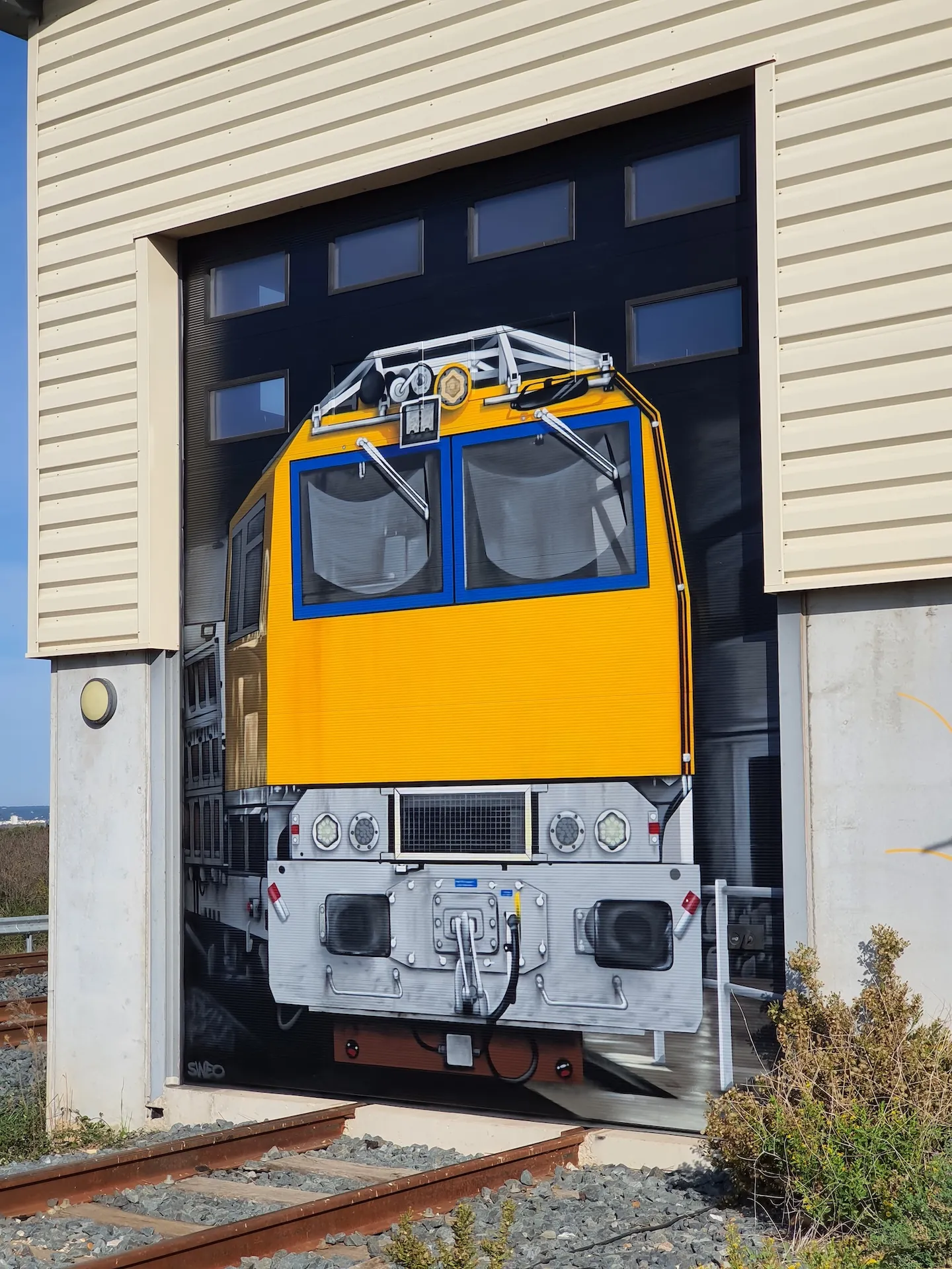 Détail d’une porte de bâtiment peinte avec l’avant d’un tramway jaune et gris, intégré à l’architecture du lieu