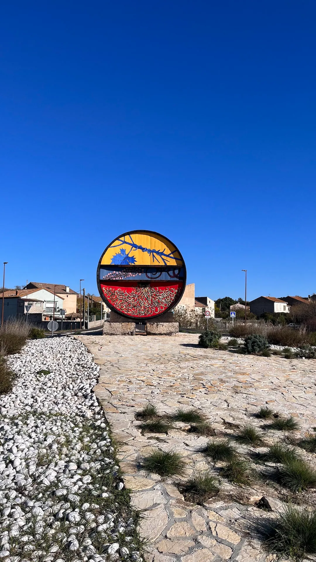 Vue éloignée d’une œuvre d’art circulaire peinte, implantée dans un paysage aride avec végétation basse et ciel bleu