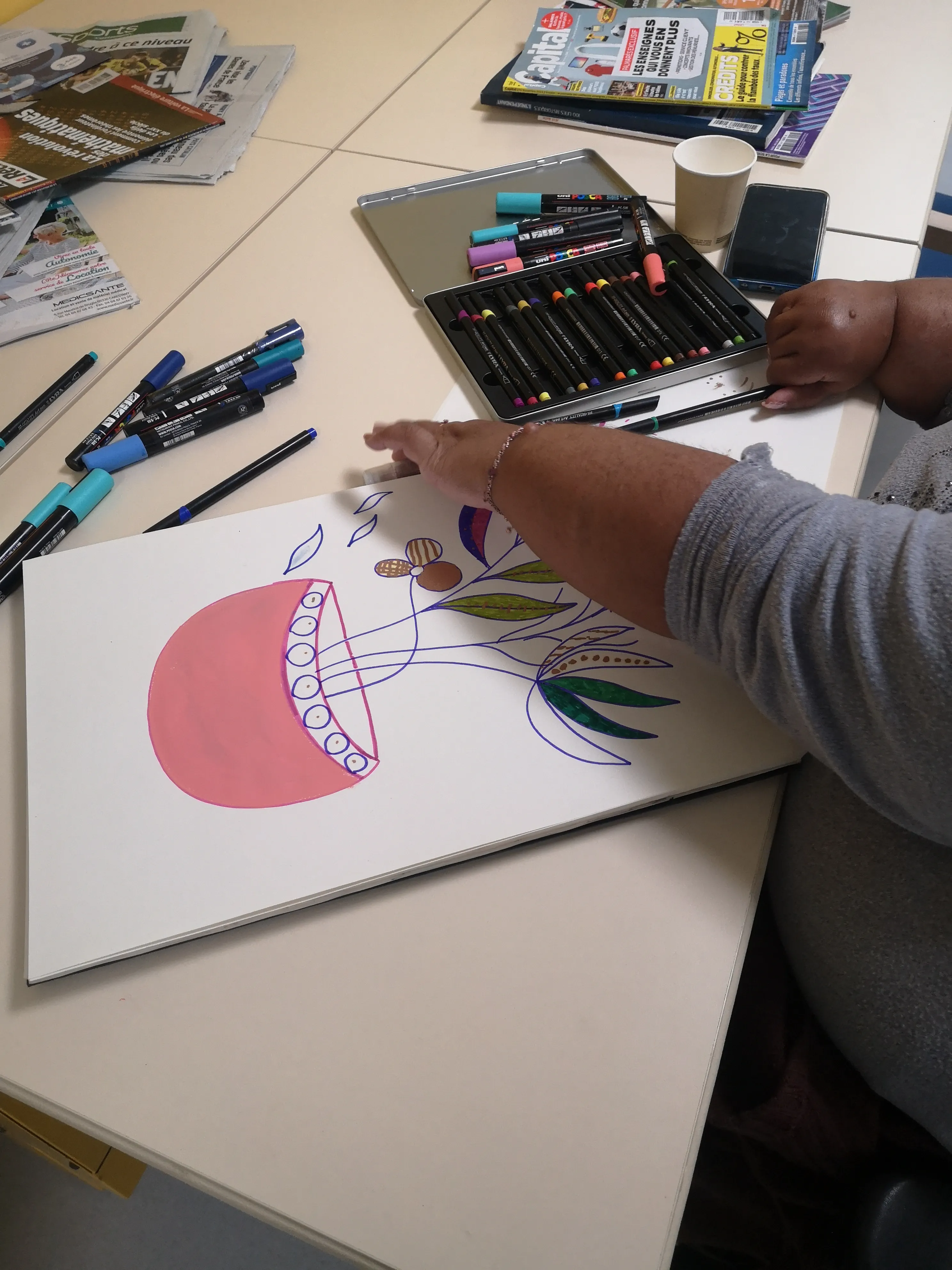 Main d’une personne dessinant une composition colorée sur papier, avec pinceaux et matériel de peinture