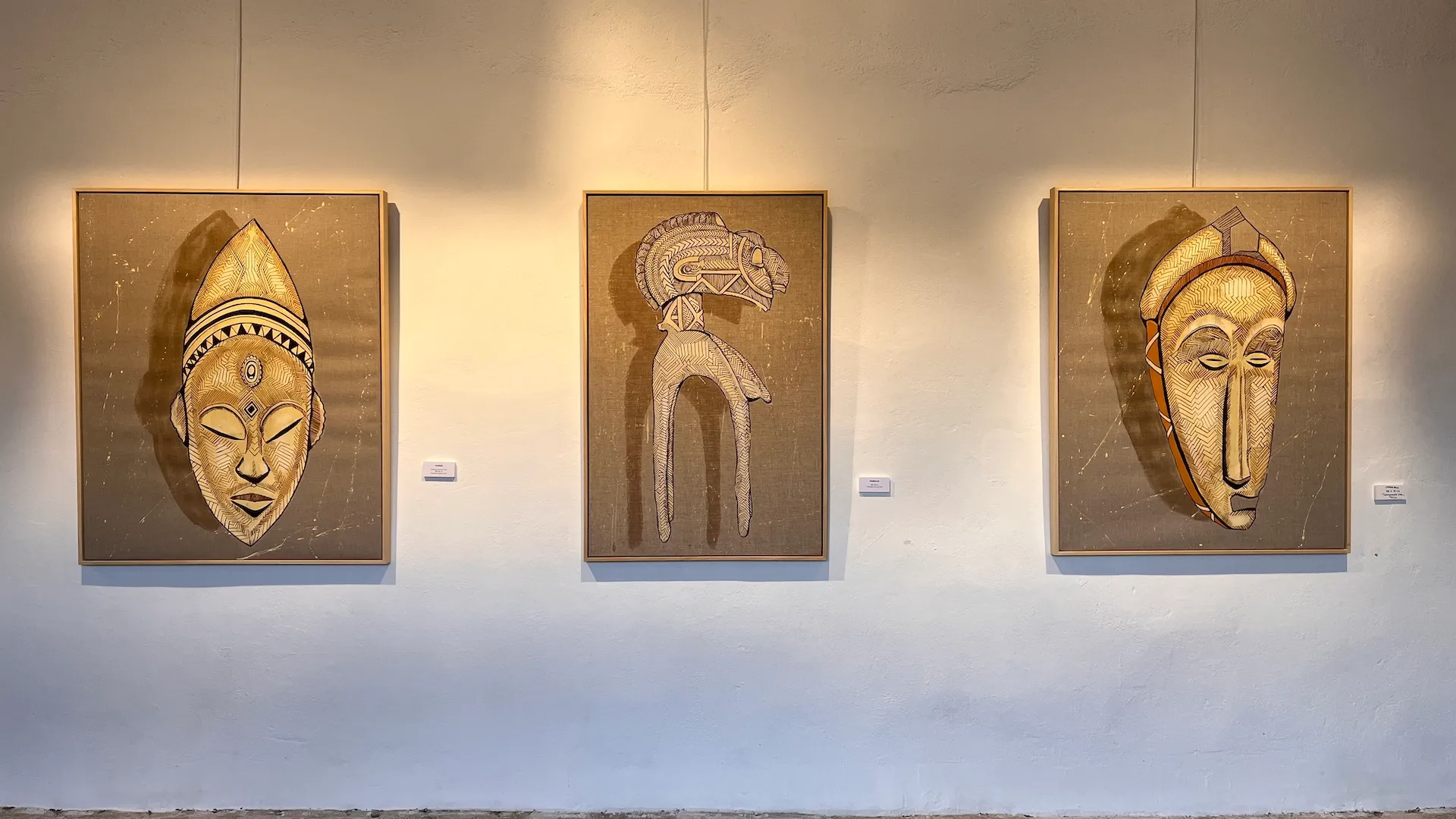 Triptyque de masques peints exposés sur un mur de galerie