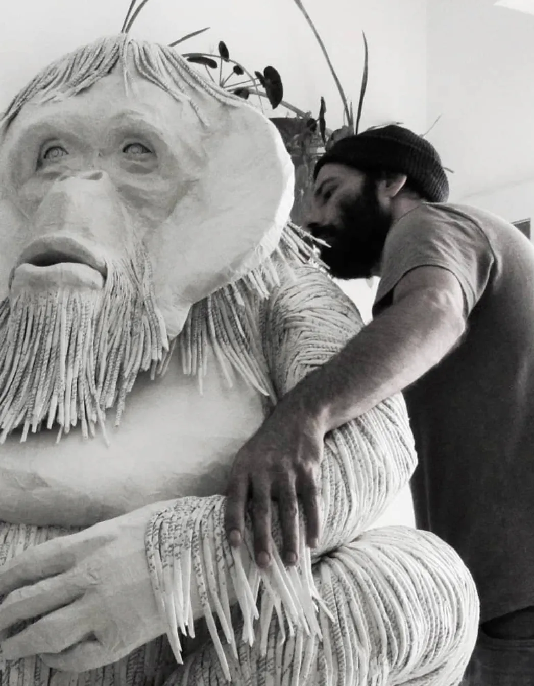 Photo en noir et blanc, un homme à droite de l'image encercle avec ses bras une sculpture de singe blanc.