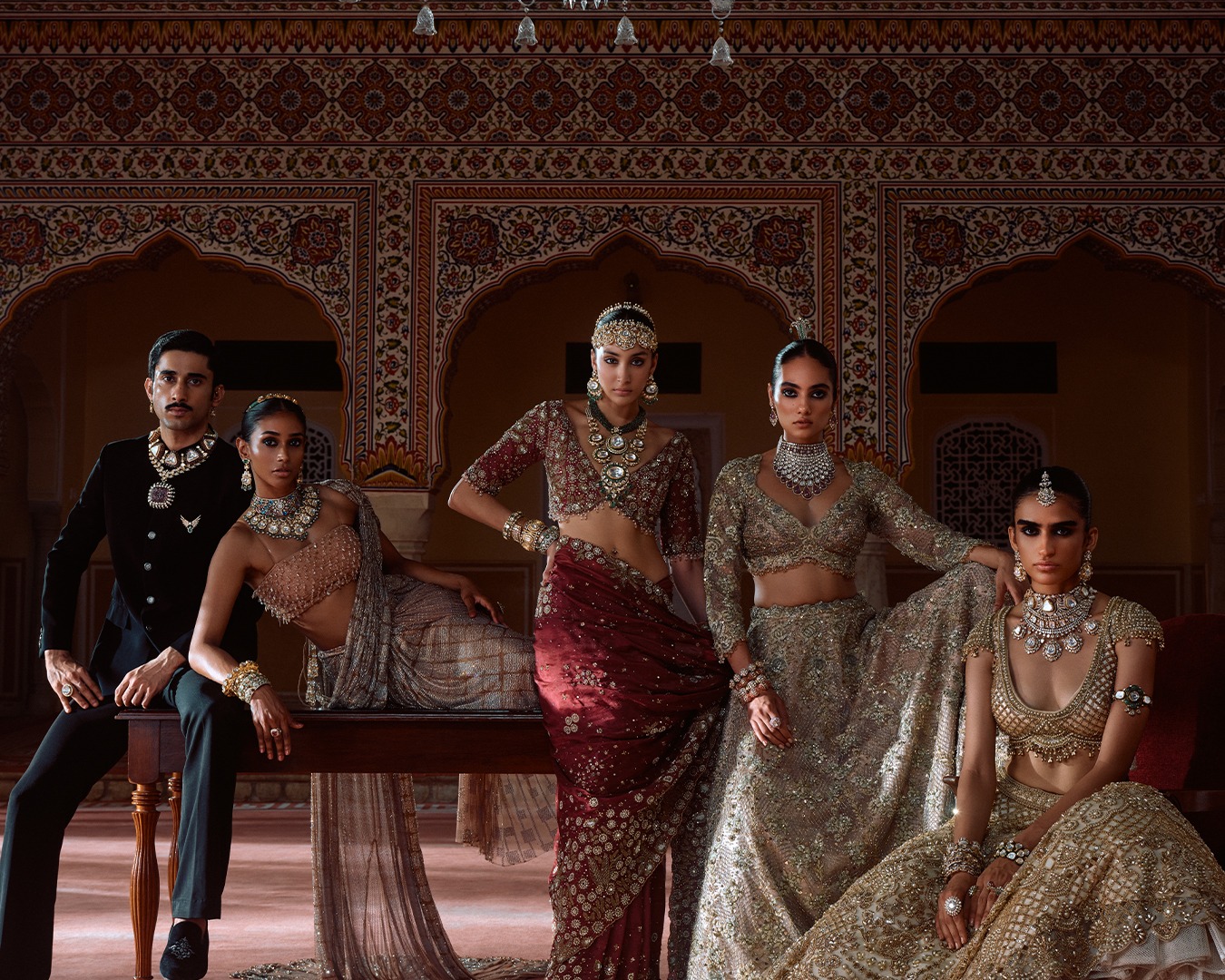 models in embroidered lehengas and a black bandhgala wear statement necklaces, maang tikkas and bangles before painted palace arches