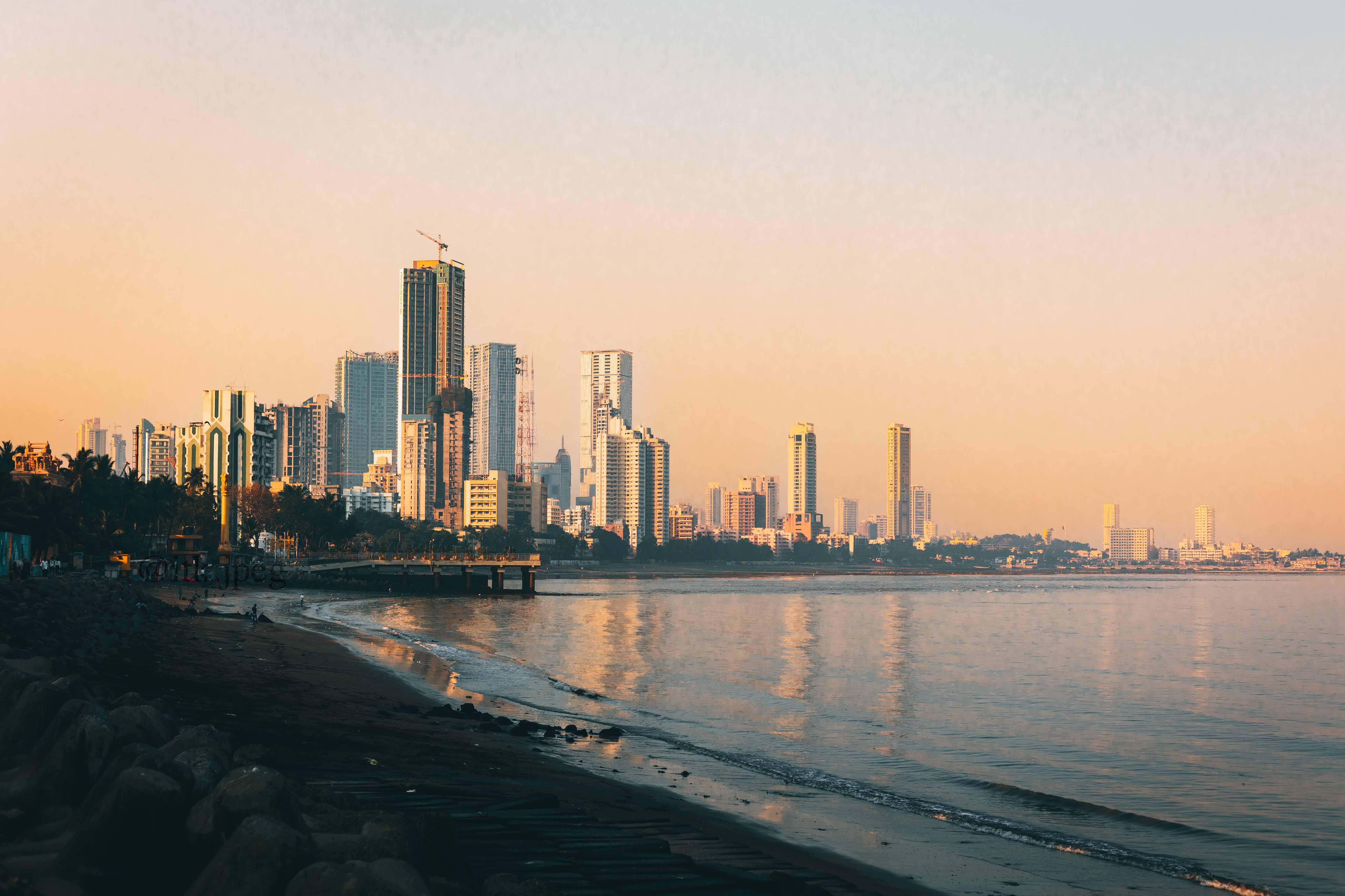City skyline at sunset with tall buildings reflecting on calm water beside a rocky beach.