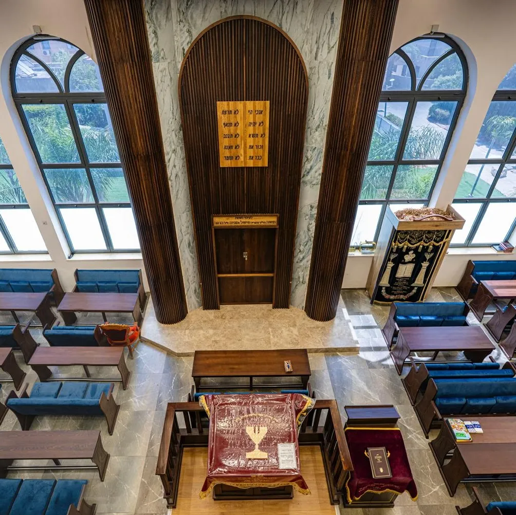 Interior of a synagogue with wooden benches, blue cushions, arched windows, and an ark at the front with Hebrew text above.