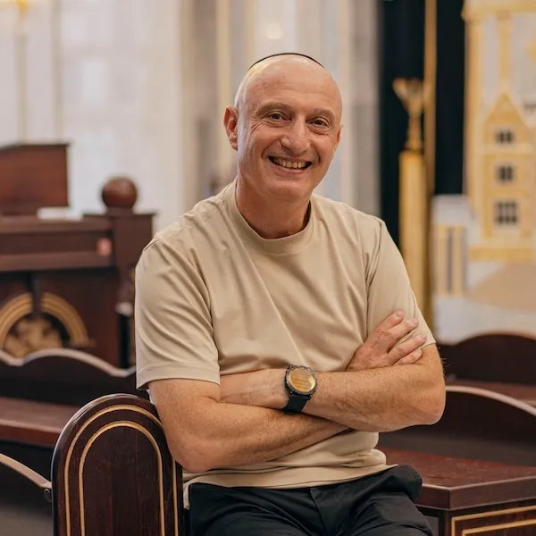 Smiling bald man wearing a beige t-shirt and black watch sitting with arms crossed on a wooden bench indoors.