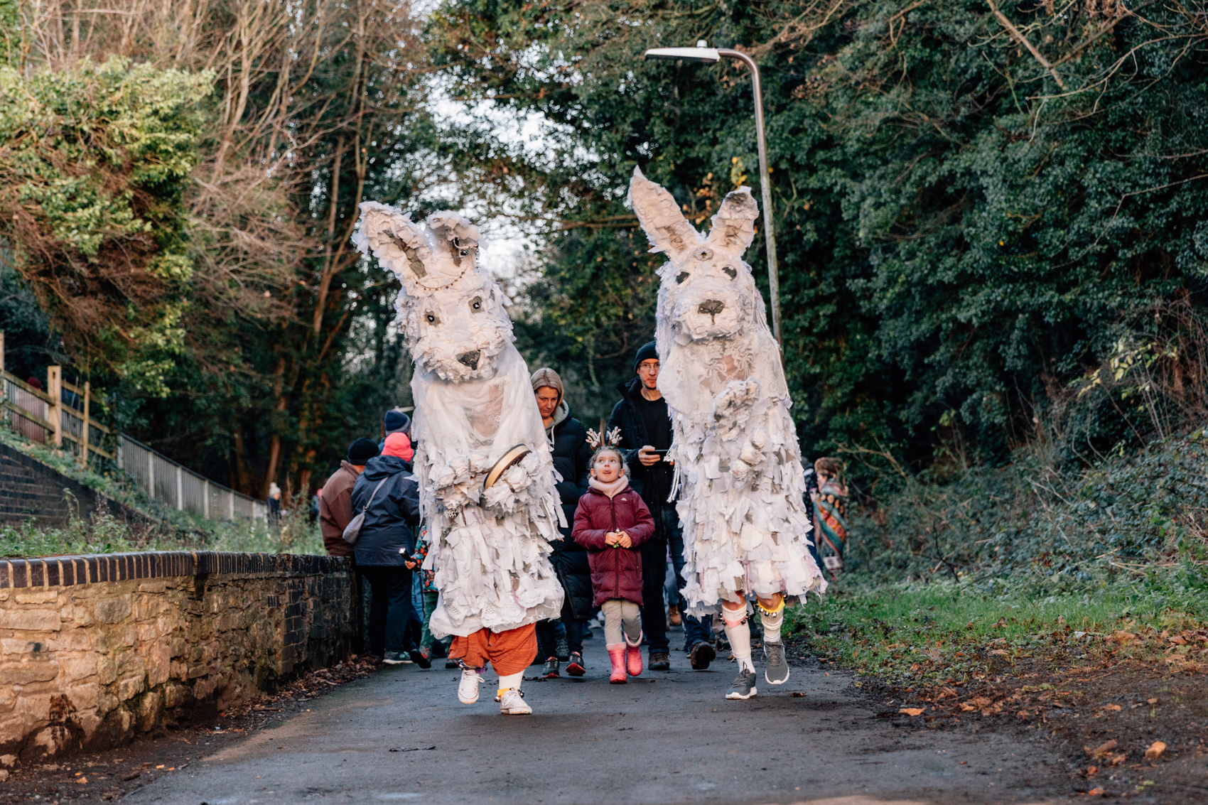 Two people in large white rabbit costumes walk with a child and adults on a wooded path.