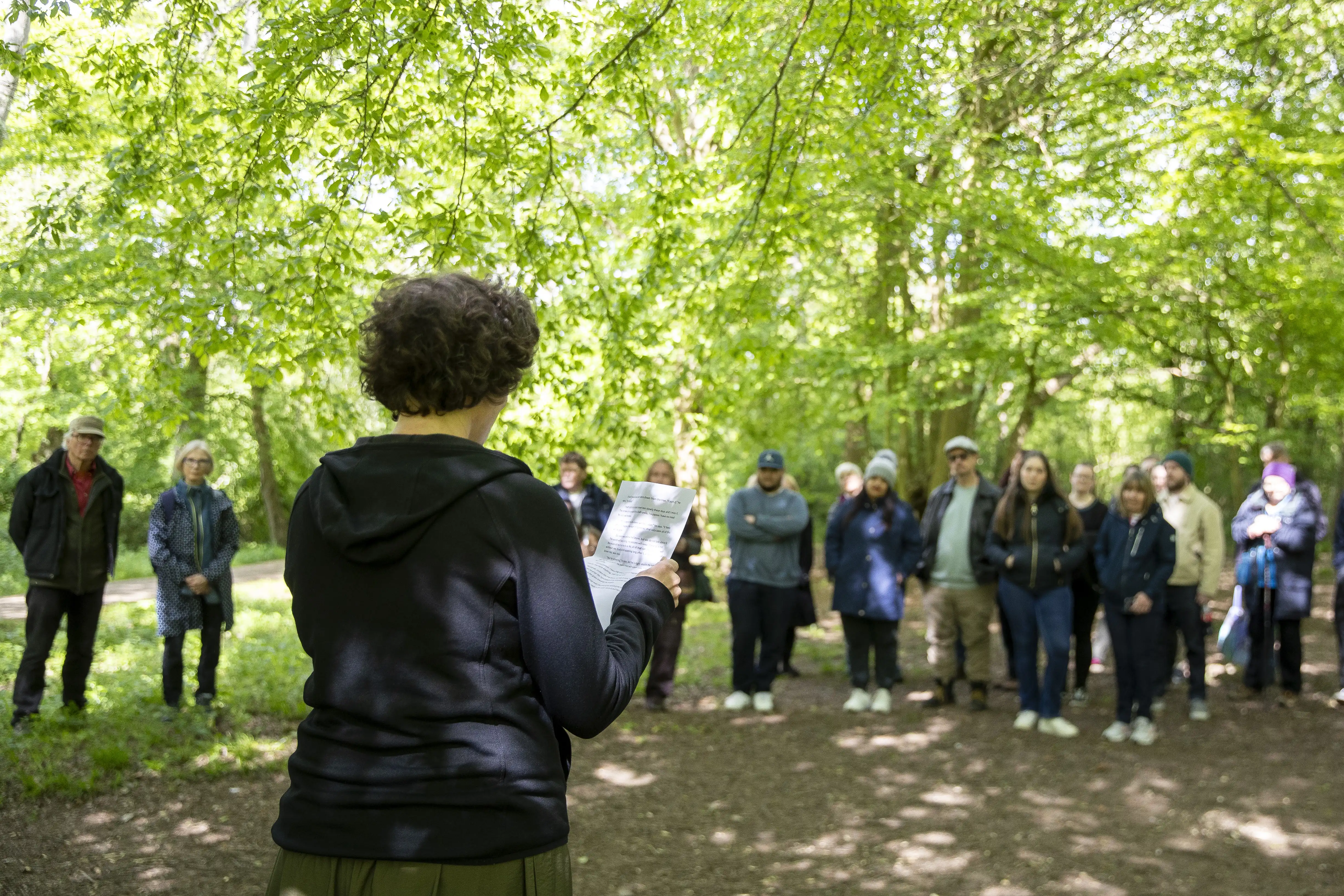 Person reading from a paper to a group standing on a forest path surrounded by green leafy trees.