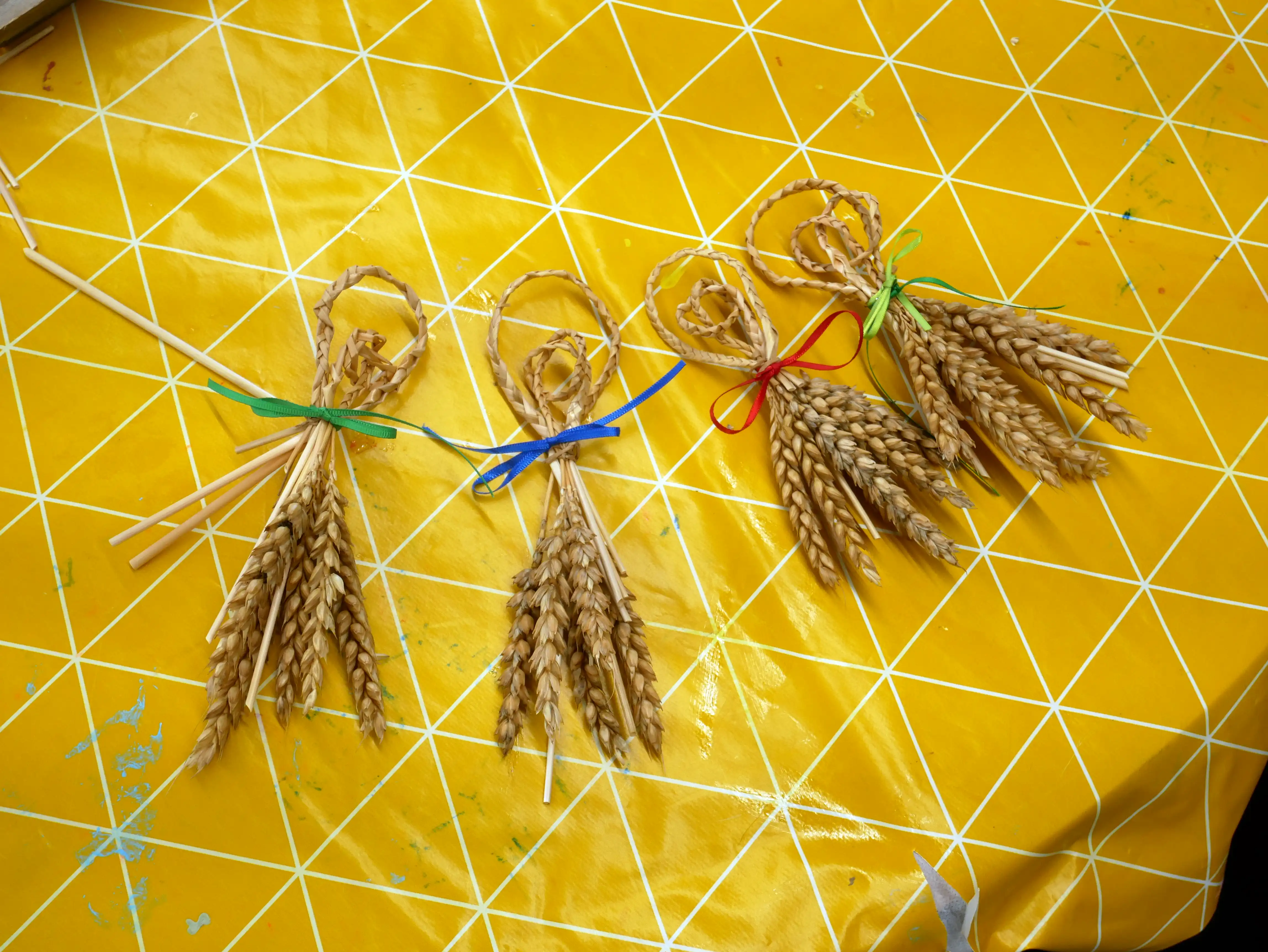 Four corn dollies made from braided wheat tied with colored ribbons on a yellow tablecloth.