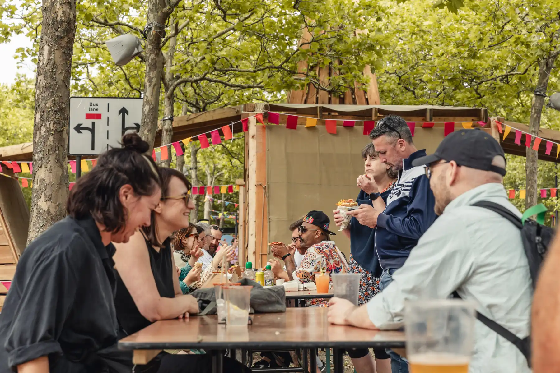 People sitting and standing around tables outdoors, eating and enjoying a festive gathering with colorful flags.