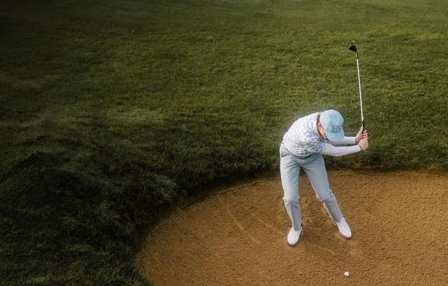 Golfer in light blue pants and cap swinging a club in a sand bunker on a golf course.