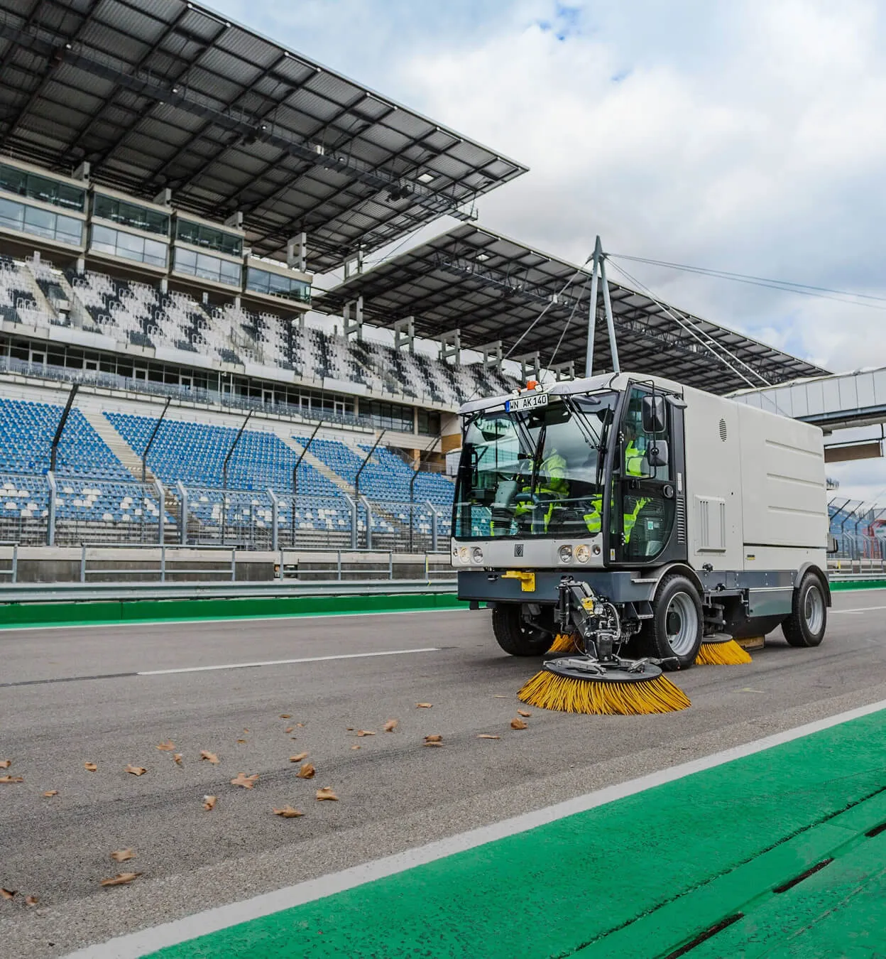 Street sweeper vehicle cleaning a paved road near empty stadium seating with blue and white seats.