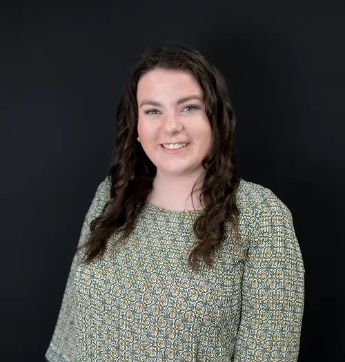 A woman with long, wavy hair smiles warmly while wearing a patterned blouse against a dark background.
