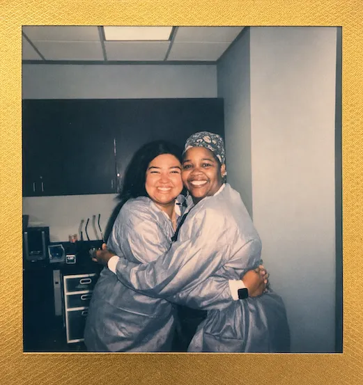 Two team members in surgical gowns hugging and laughing in the office, polaroid-style photo