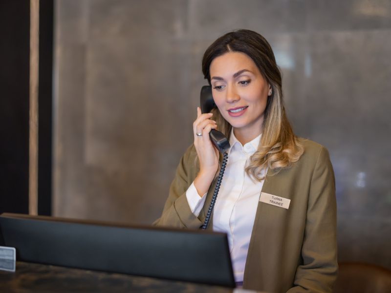 Hotel front desk employee speaking on a desk phone at the reception counter.