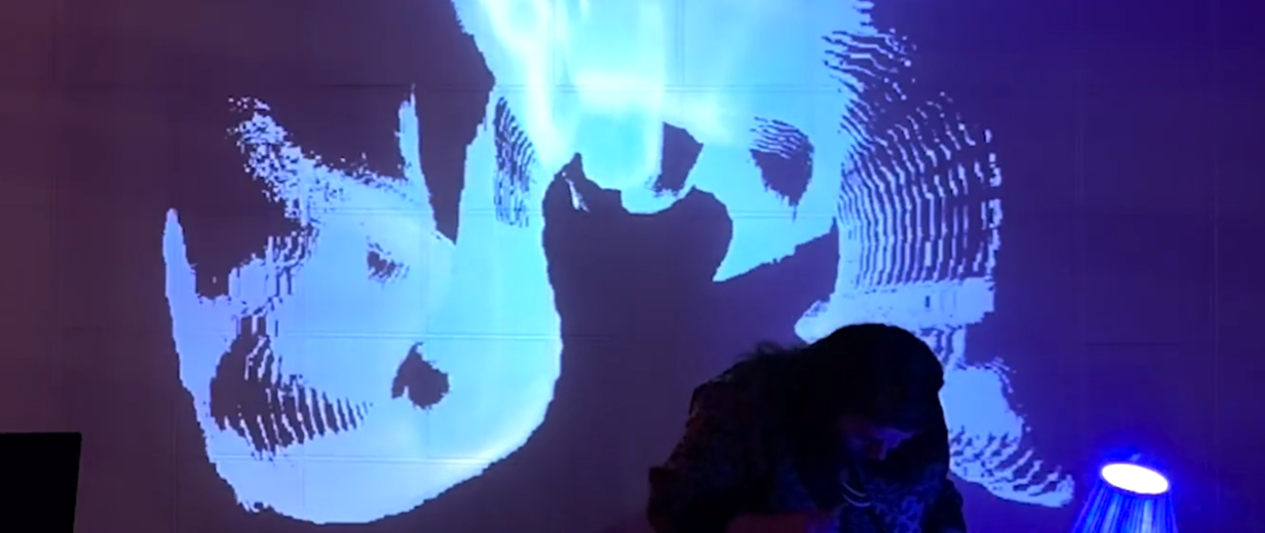 A woman bends over a table with a small lamp in a darkened room. On the wall behind her are projected bright flowing lines like brush strokes, rippling and vibrating across the wall. 