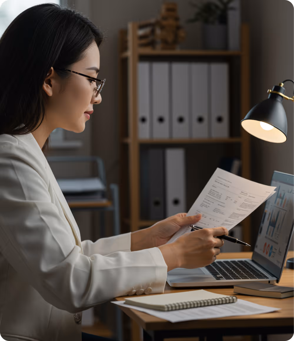 Woman in glasses reviewing paperwork while sitting at a desk with a laptop, notebook, and a desk lamp.
