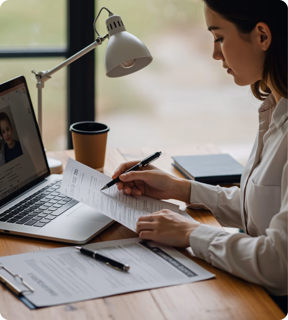 Woman reviewing and marking paperwork at a wooden desk with a laptop showing a child's photo, a desk lamp, and a coffee cup nearby.