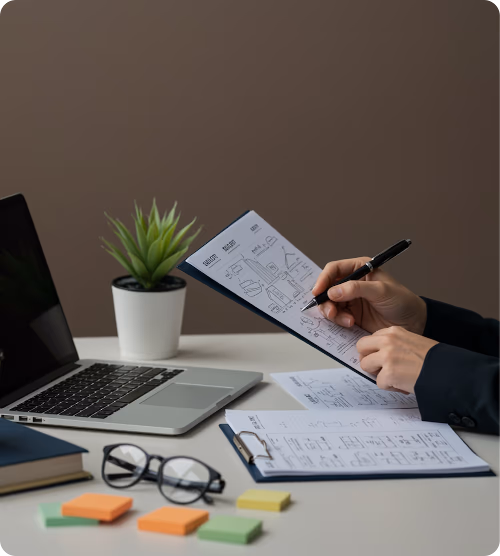 Person holding a clipboard and annotating a document with sketches and notes at a desk with a laptop, eyeglasses, sticky notes, and a potted plant.