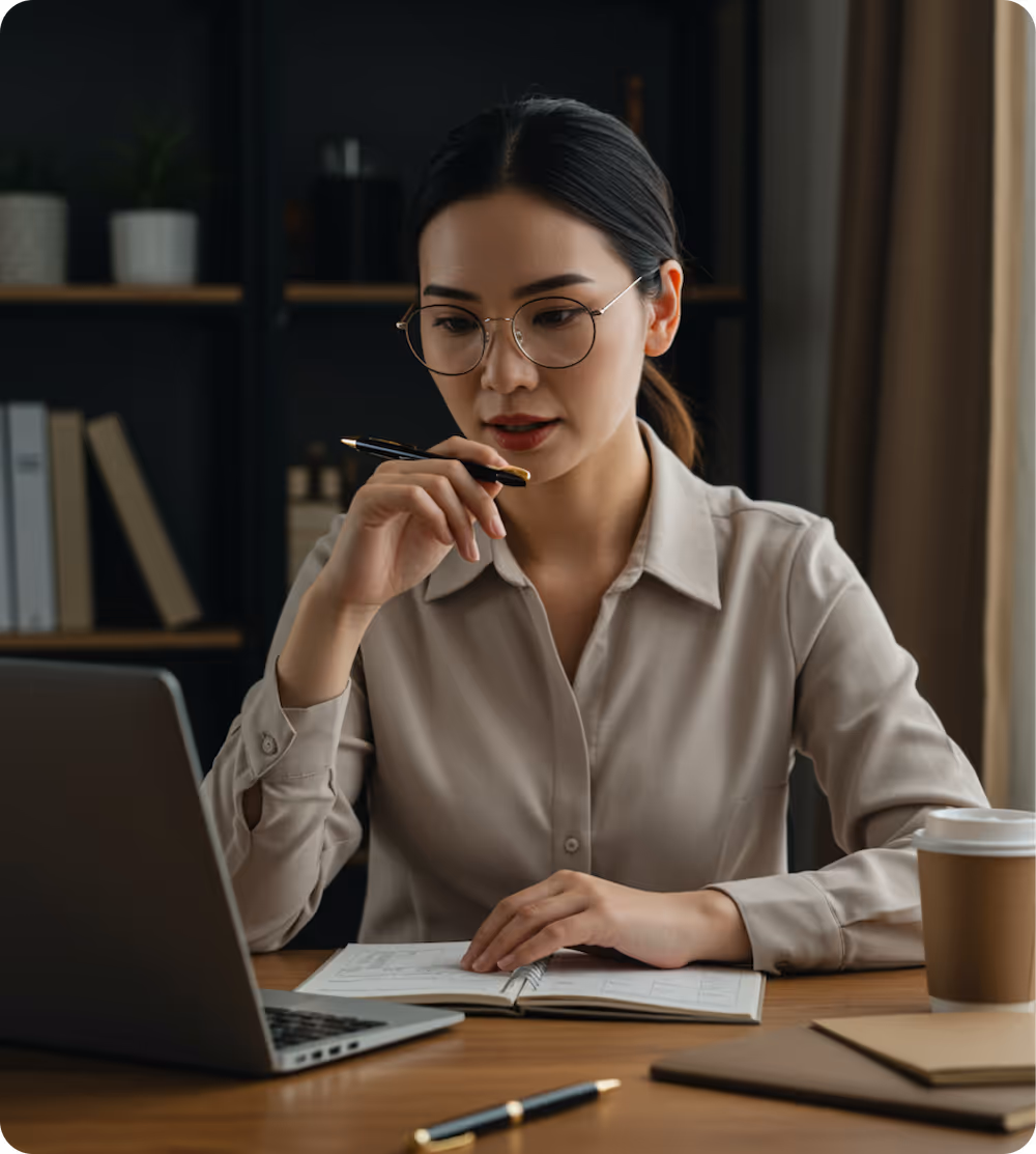 Woman wearing glasses thoughtfully looking at a laptop with an open planner on a wooden desk, holding a pen near her mouth.