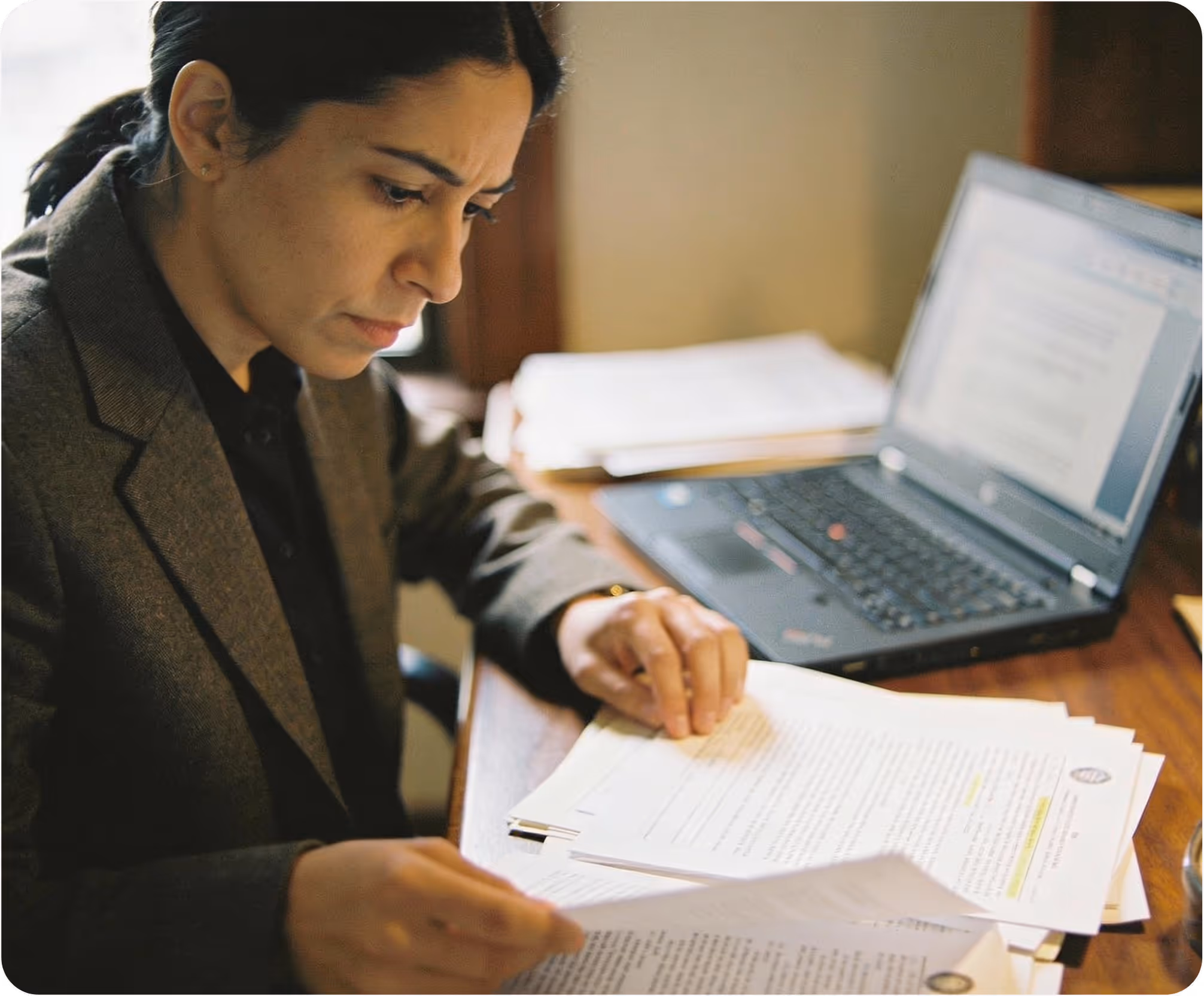 Woman in a blazer carefully reviewing legal documents at a desk with a laptop.