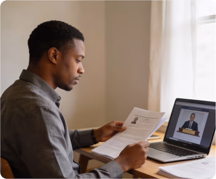 Man seated at desk reading documents with a laptop open showing a man speaking at a podium.