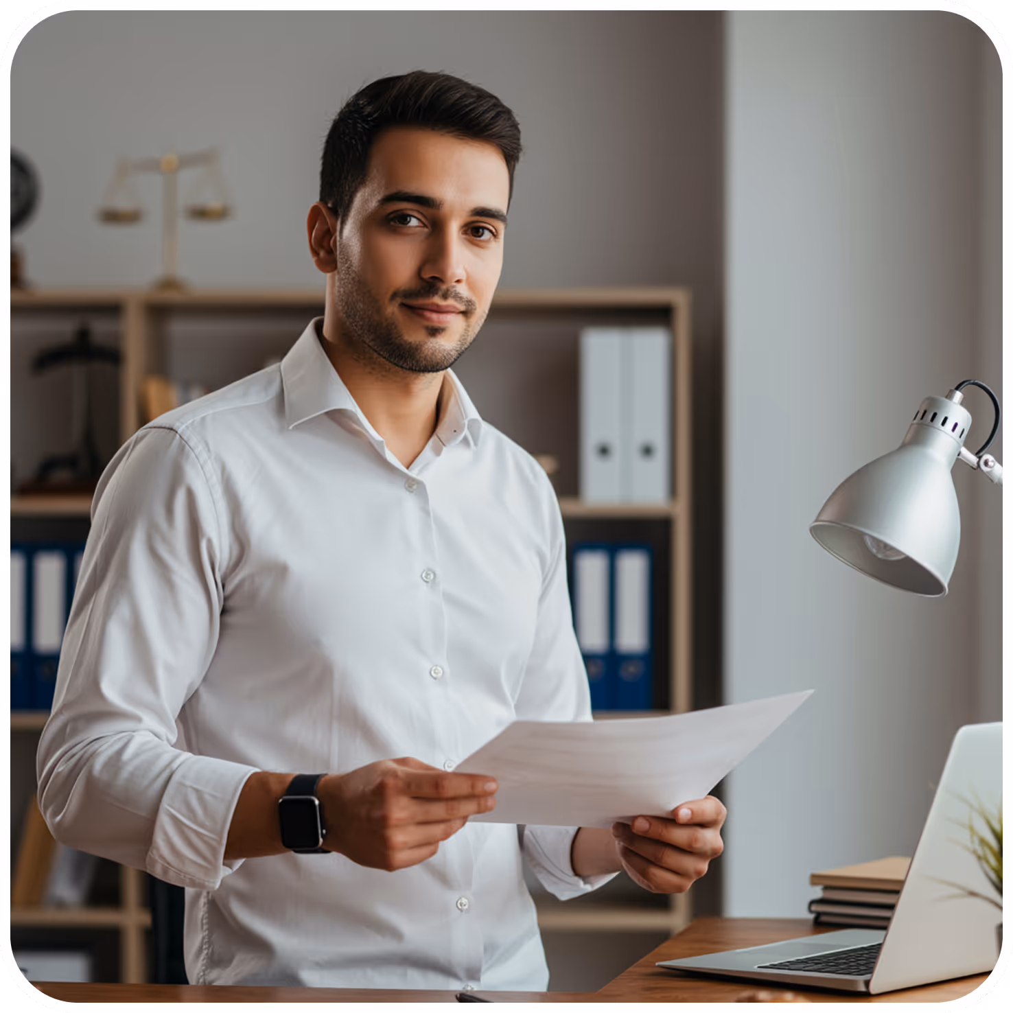 Man in a white shirt holding papers in an office setting with a laptop and desk lamp nearby.