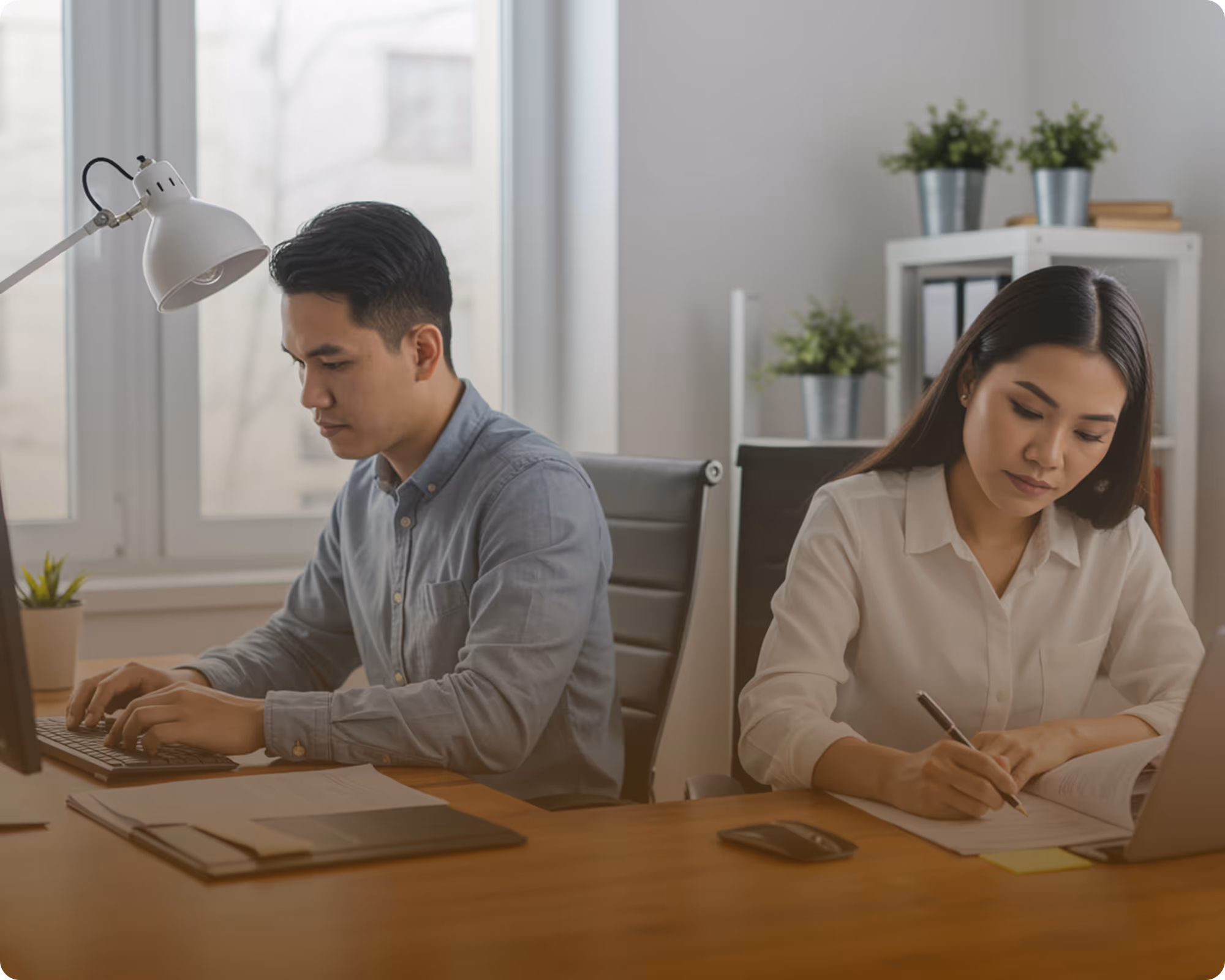 Two focused professionals working side by side at a desk, one typing on a keyboard and the other writing in a notebook.