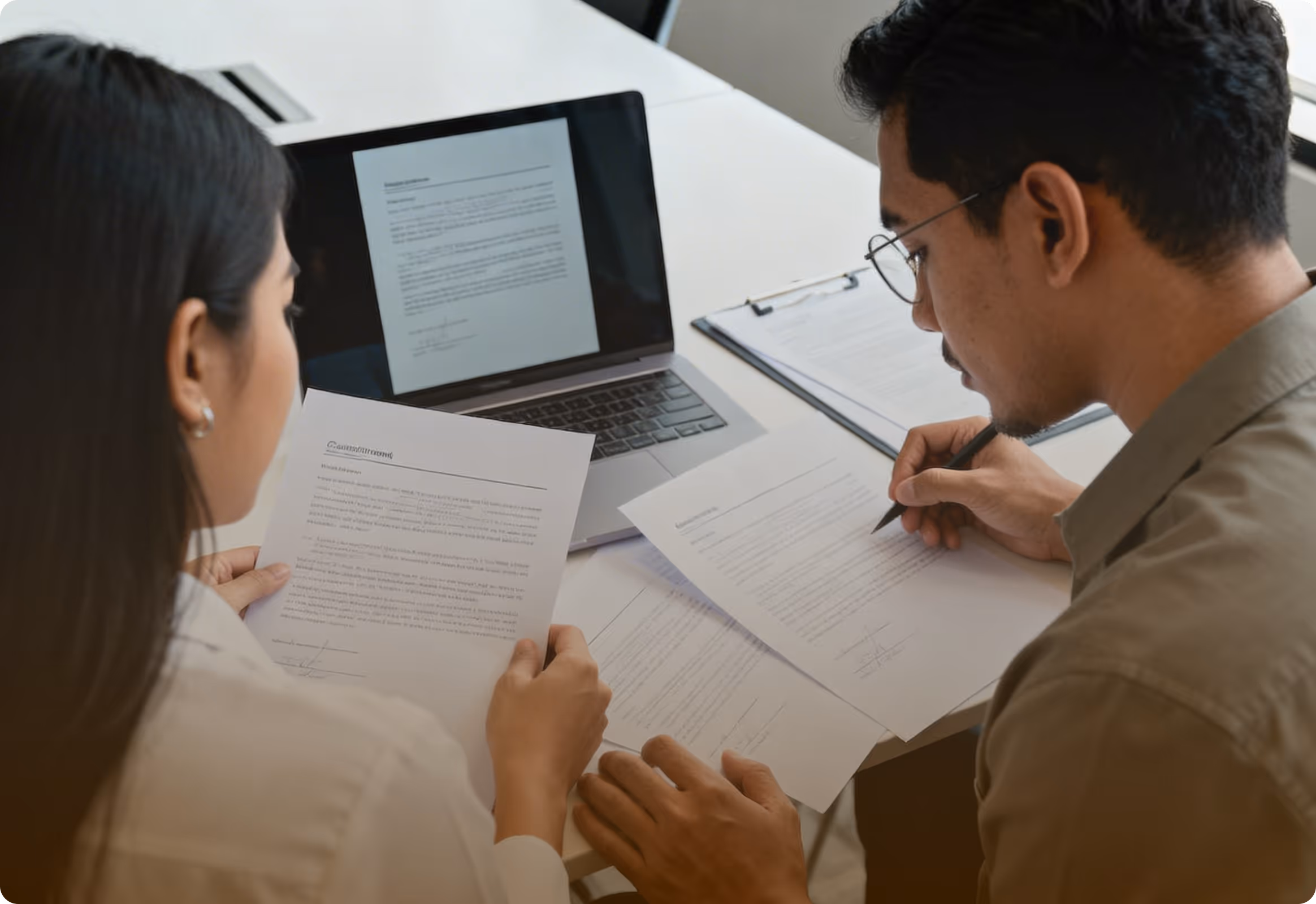 Two people reviewing and signing documents at a desk with a laptop and clipboard.