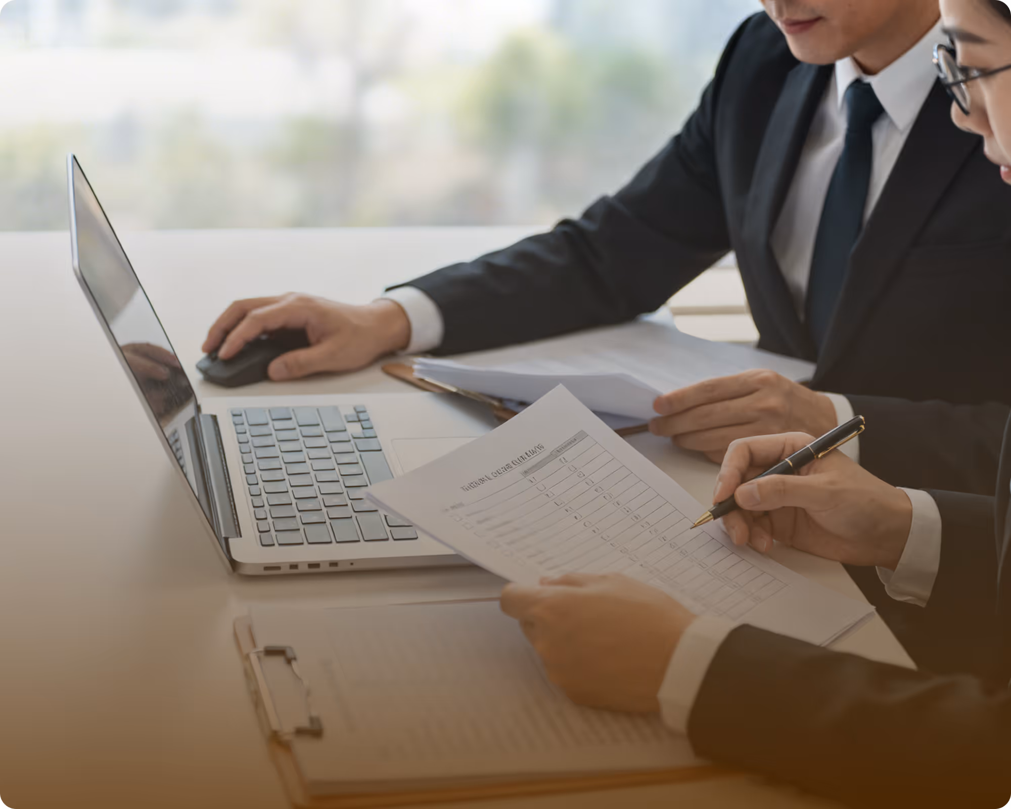 Two professionals in suits reviewing documents and using a laptop at a desk.