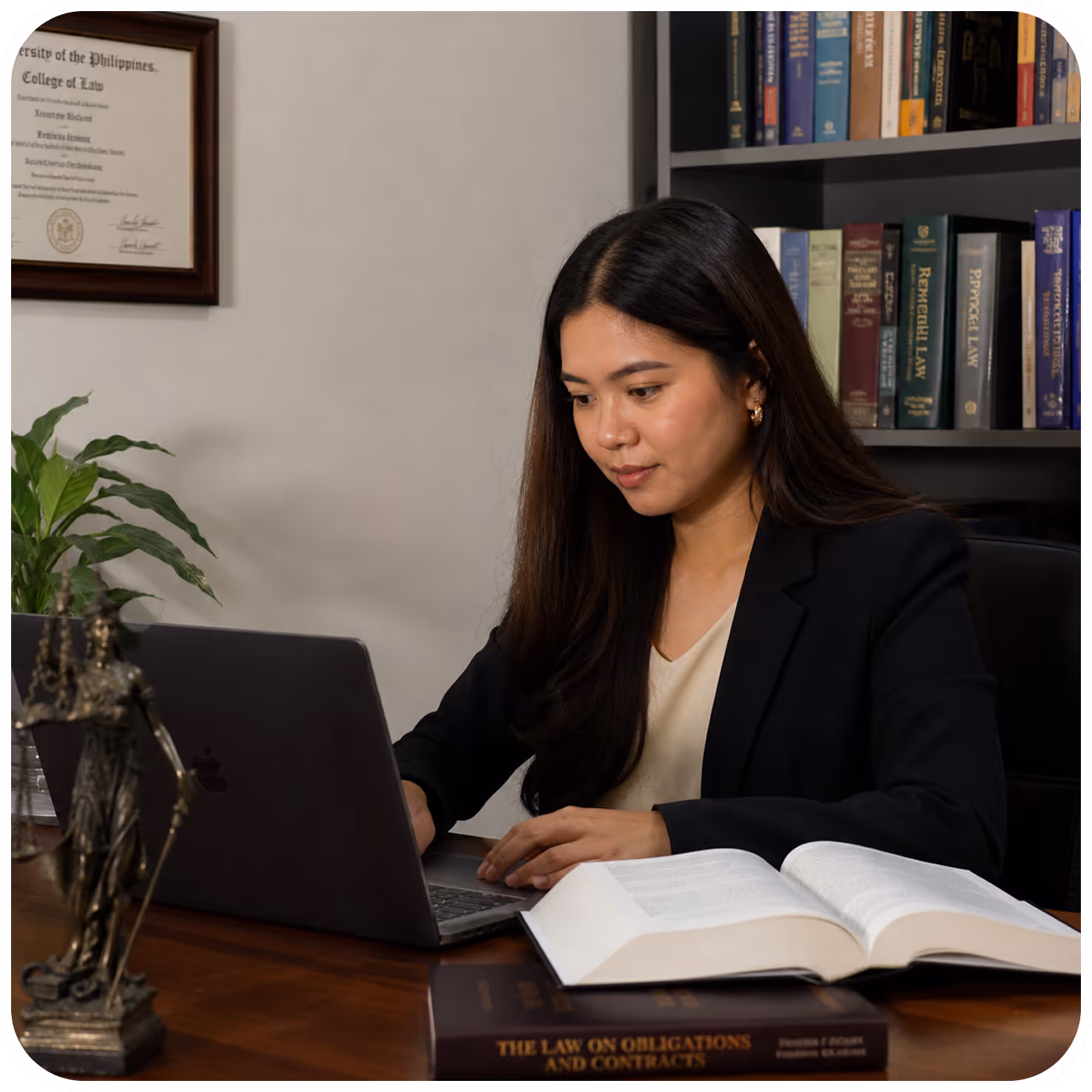 Woman in a black blazer working on a laptop at a desk with an open law book and bookshelf in the background.
