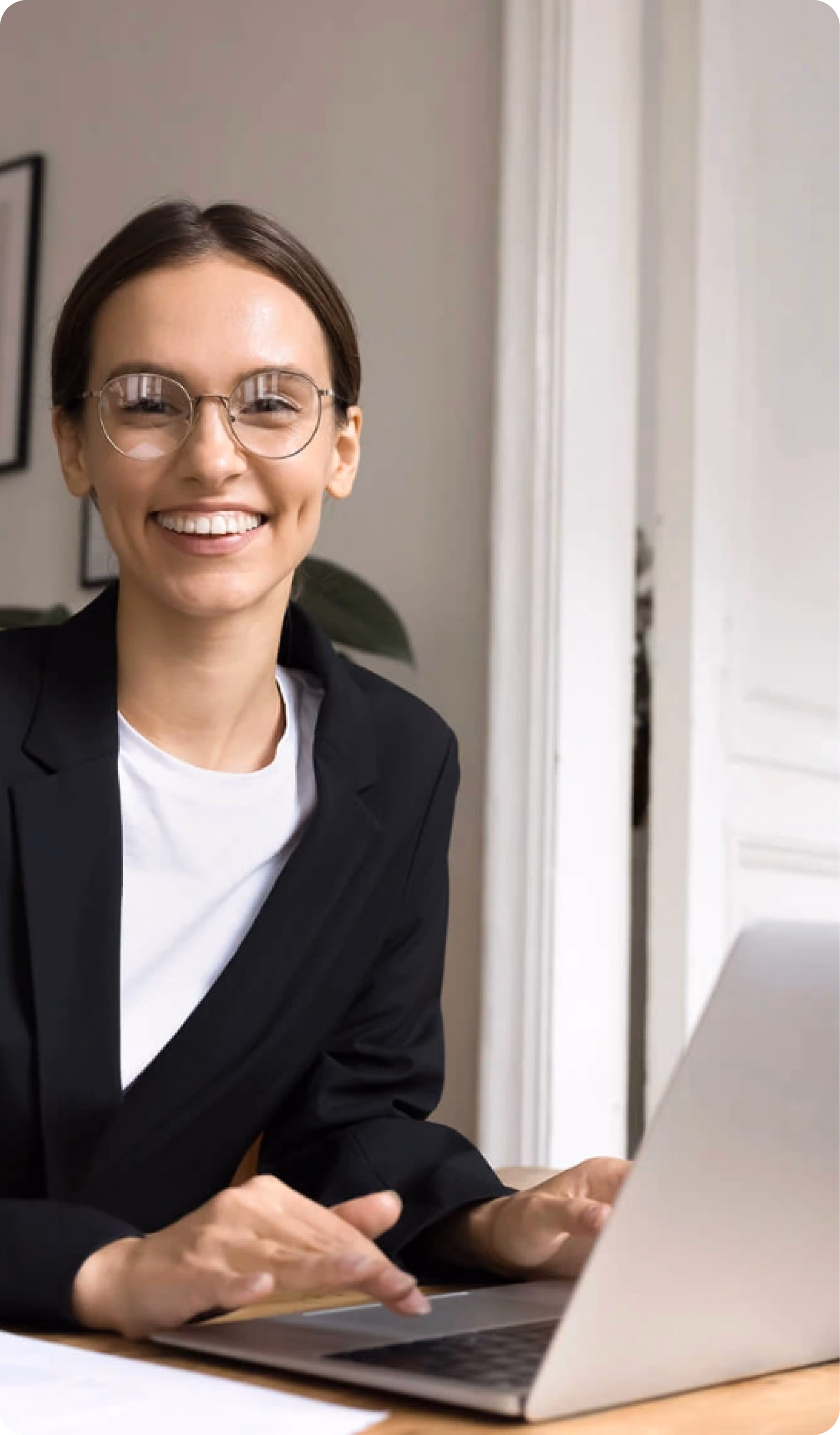Smiling young woman with glasses in black blazer using a laptop at a desk indoors.