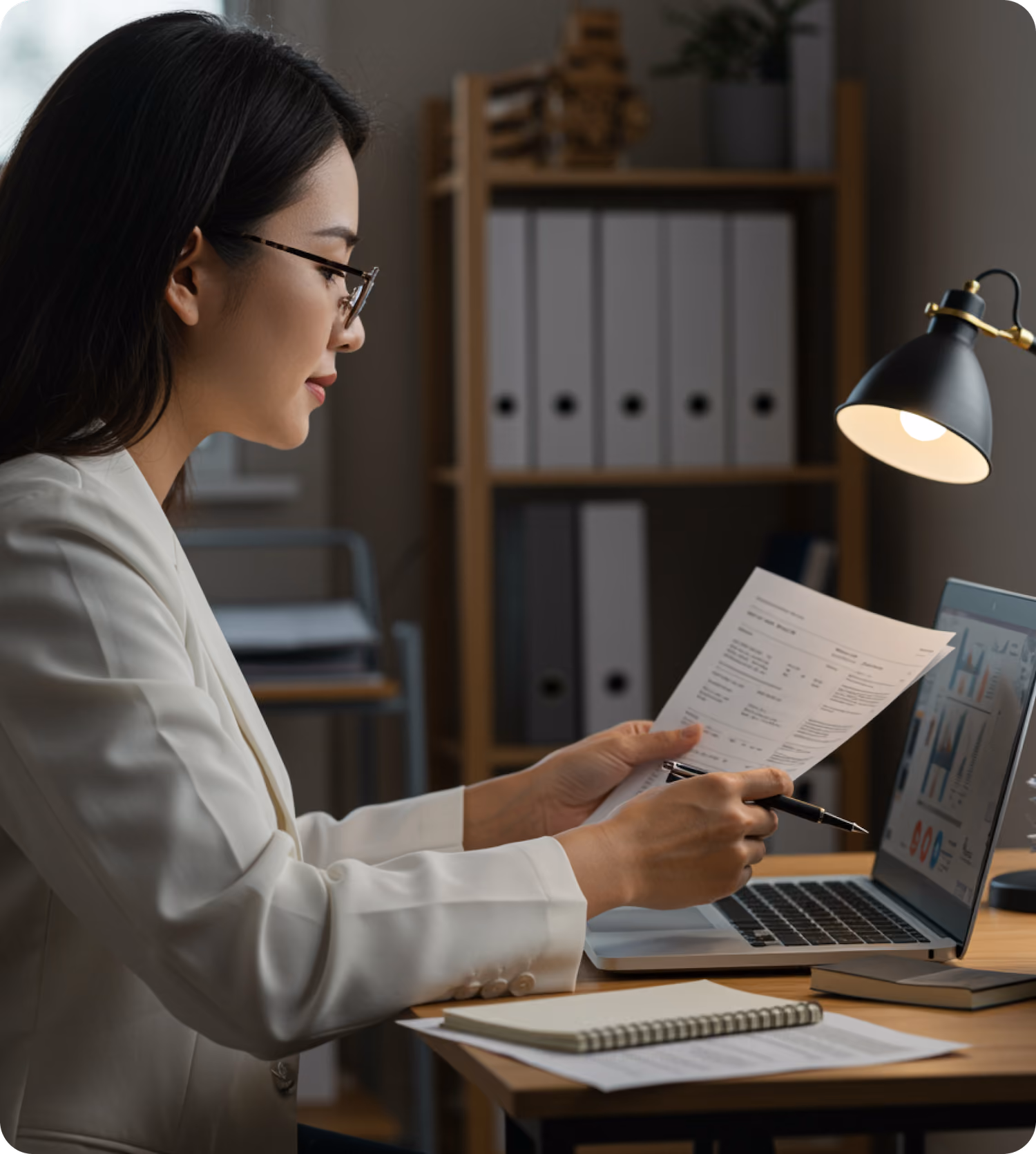 Woman in glasses and white blazer reviews documents while working on a laptop at a desk with a lamp.