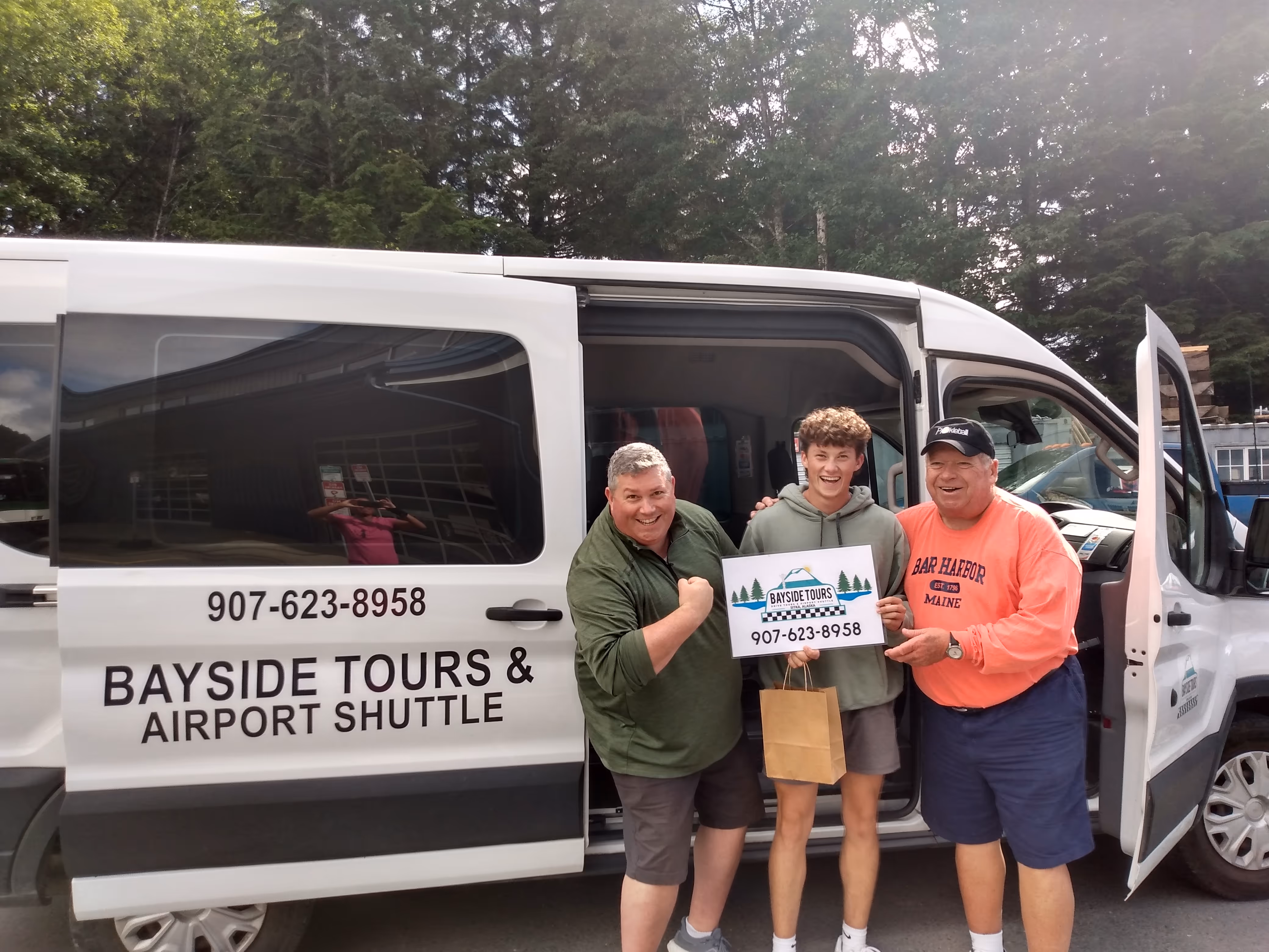 Three men smiling in front of a Bayside Tours & Airport Shuttle van, one holding a sign with the company name and phone number.