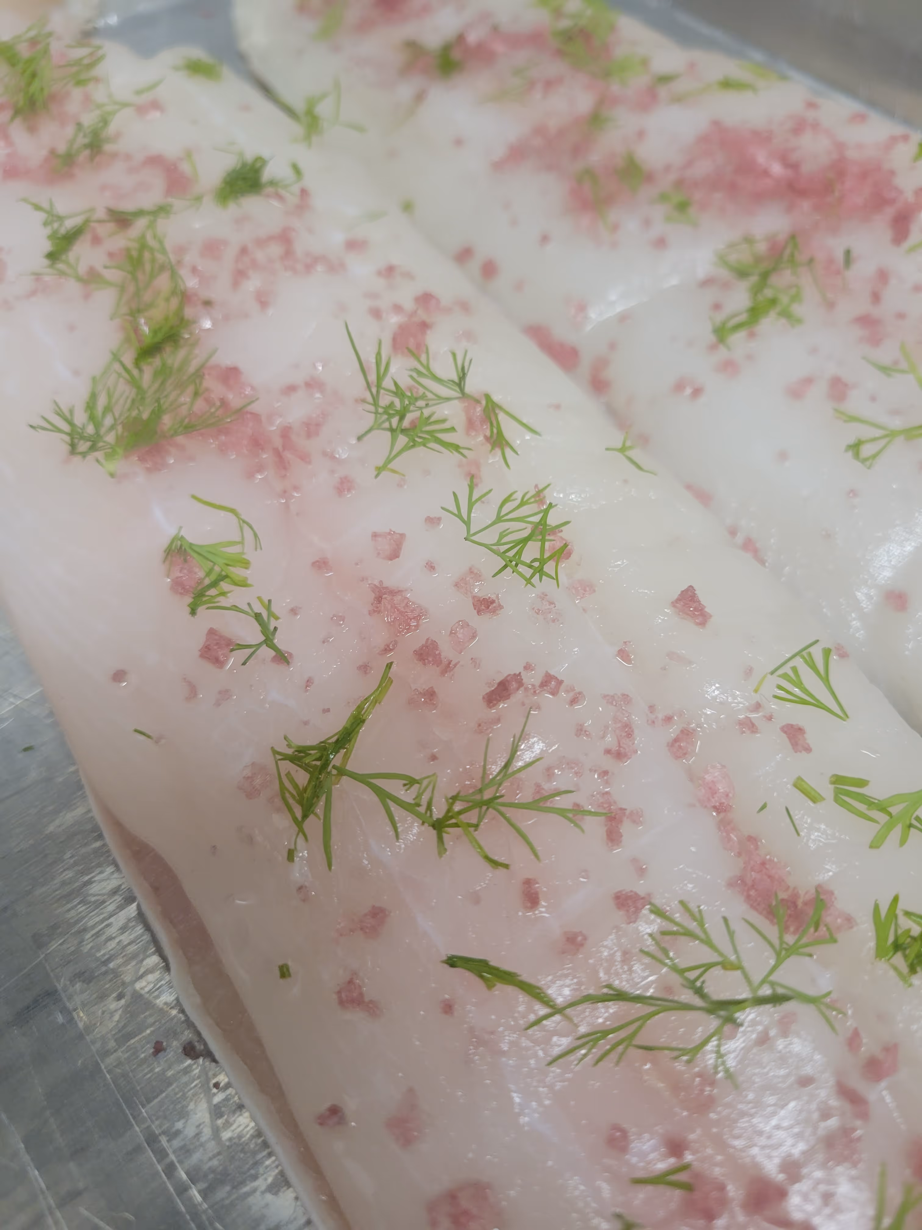 Close-up of raw white fish fillets seasoned with pink salt crystals and fresh green dill sprigs.