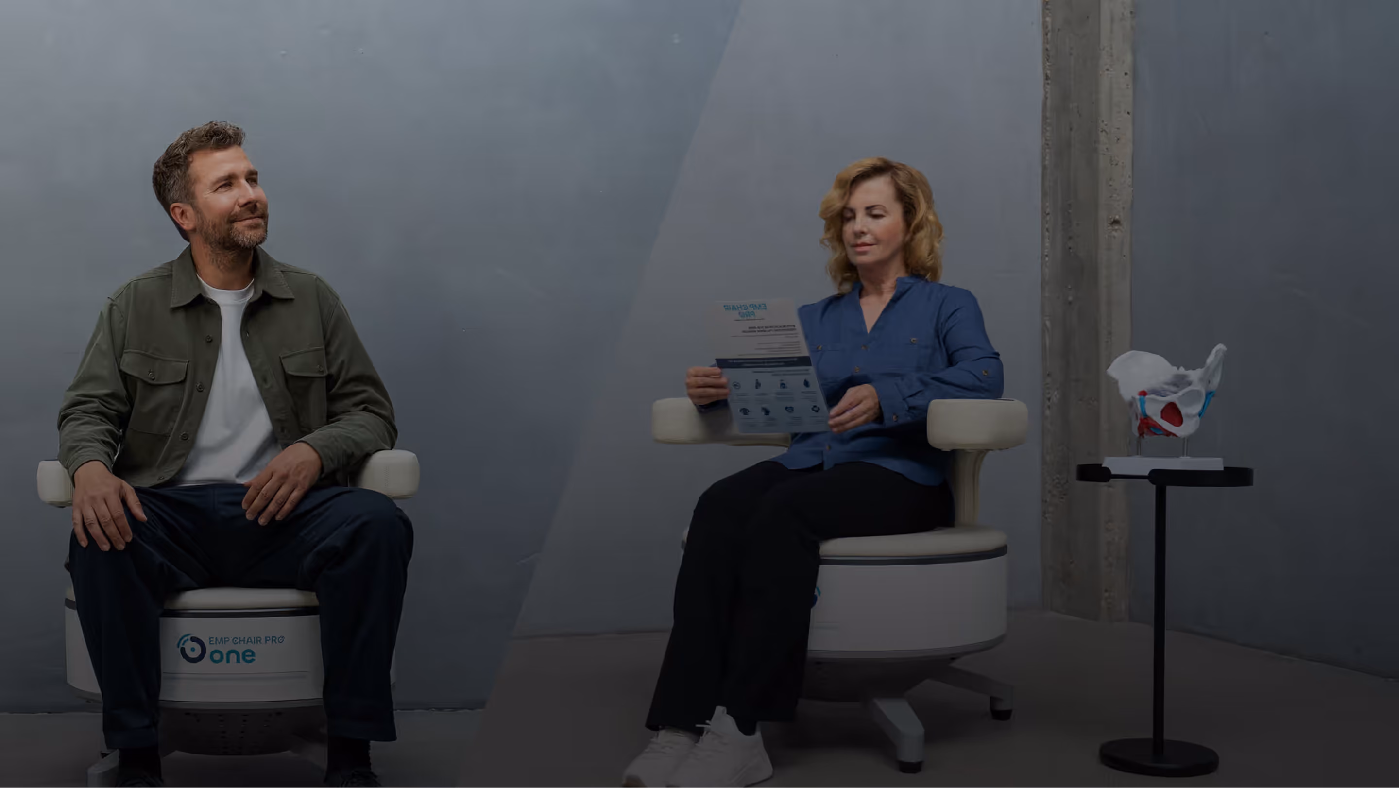 A man and a woman seated in a modern, minimalist room; the man wears a green jacket and looks left, while the woman in a blue blouse reads a pamphlet, with a pelvic bone model on a table nearby.