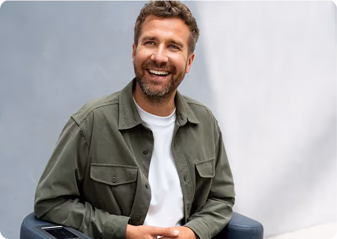 Smiling man with short brown hair and beard wearing an olive green jacket over a white shirt, sitting on a dark chair against a light background.