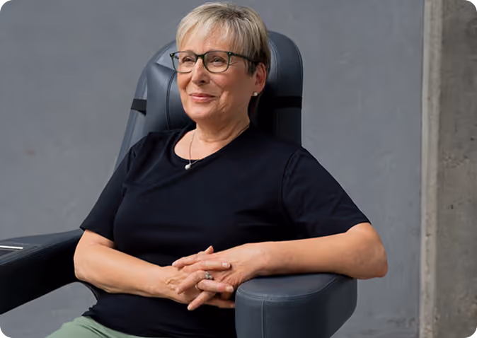 Smiling older woman with short blonde hair and glasses sitting in a gray chair with hands clasped in her lap against a plain gray background.