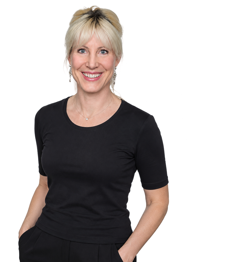 Smiling woman with blonde hair wearing a black short-sleeve shirt and black pants, standing with hands in pockets, with a white palm leaf silhouette in the background.