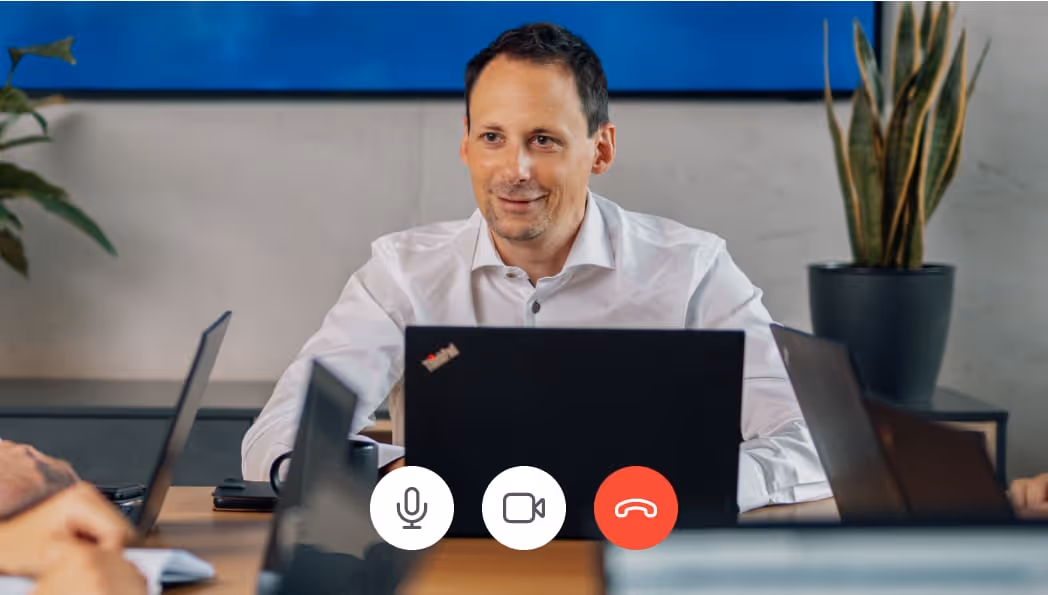 Dr. Sebastian Adam in weißem Hemd sitzt an einem Holztisch mit Laptop und nimmt an einer Videokonferenz teil.