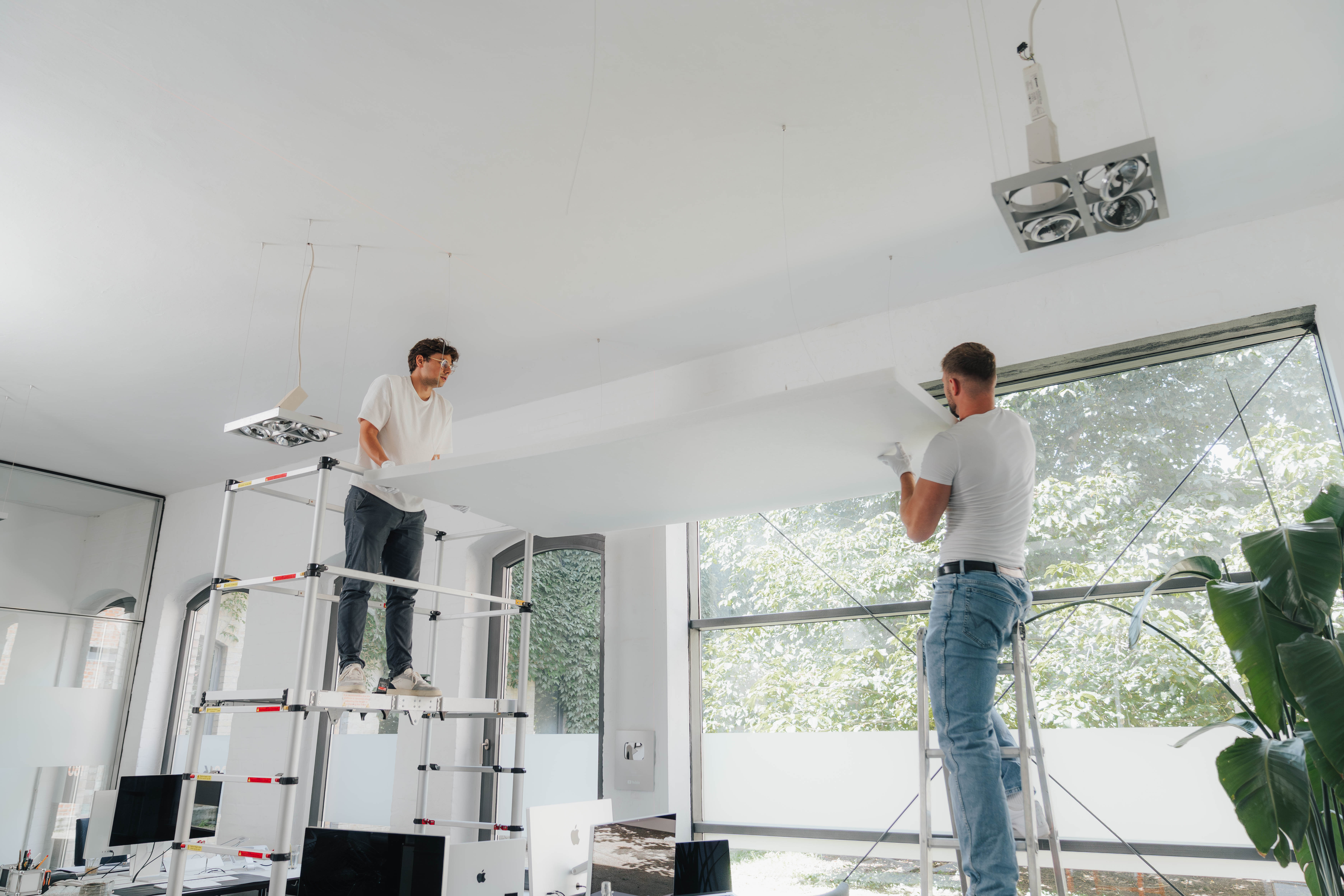 Two men installing a large ceiling panel in a bright office with large windows and computer desks below.