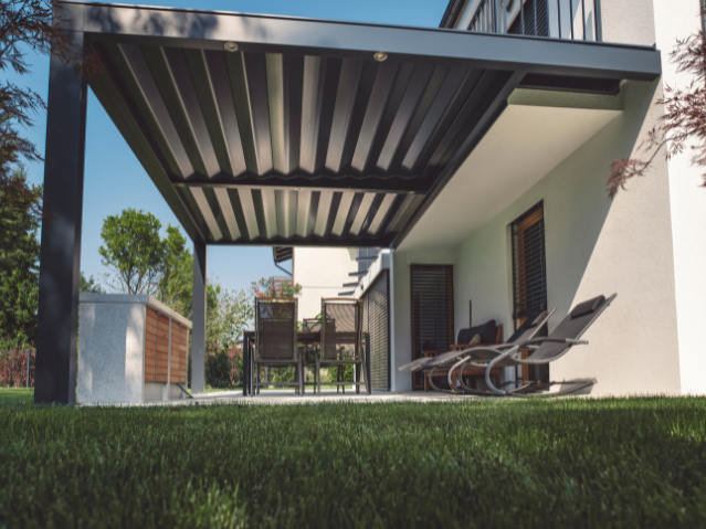 Terrasse couverte d'une maison moderne avec chaises longues à droite et une table avec plusieurs chaises sous un auvent métallique gris.