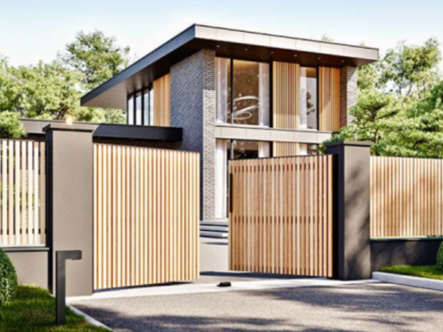 Maison moderne à deux étages avec grandes fenêtres, façade en briques et bois, entourée d'une clôture et un portail en bois clair, avec un chemin et des arbres verts autour.