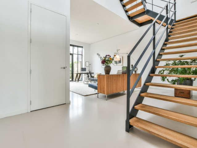 Intérieur moderne avec escalier en bois et métal à droite, meuble en bois avec vase de fleurs, grand espace lumineux avec canapé et bureau au fond.