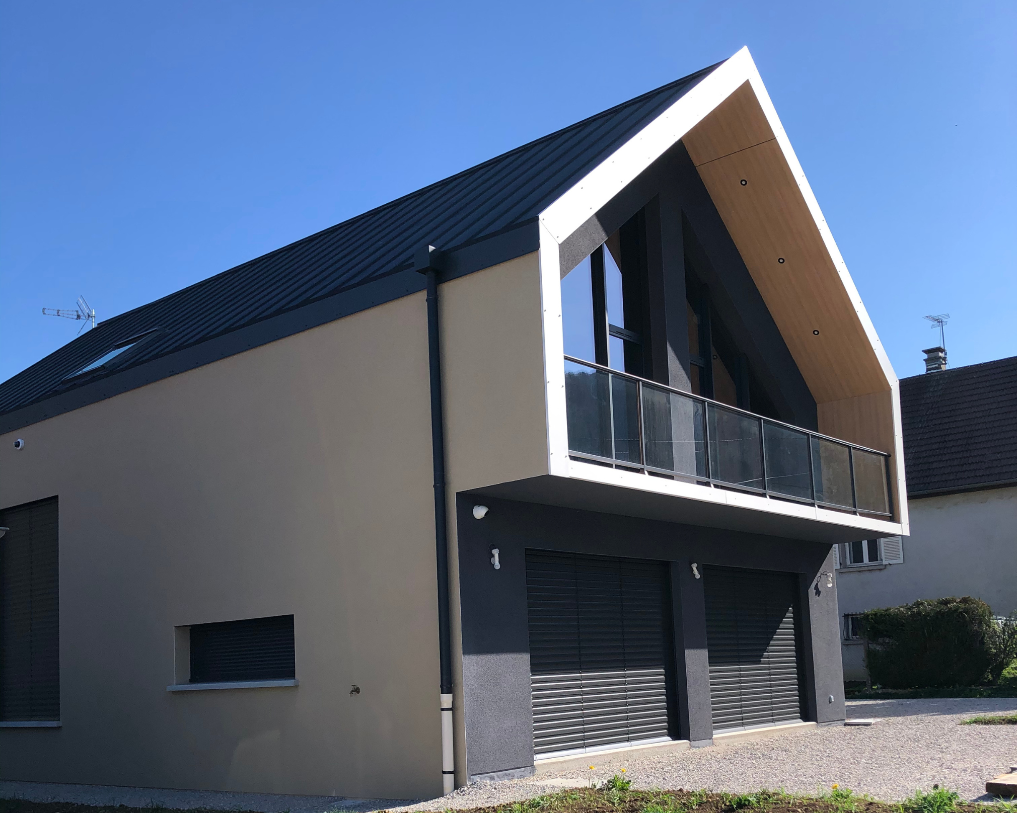 Maison moderne à deux étages avec balcon vitré, murs beige et gris, toit métallique noir incliné et grandes fenêtres sous un ciel bleu clair.