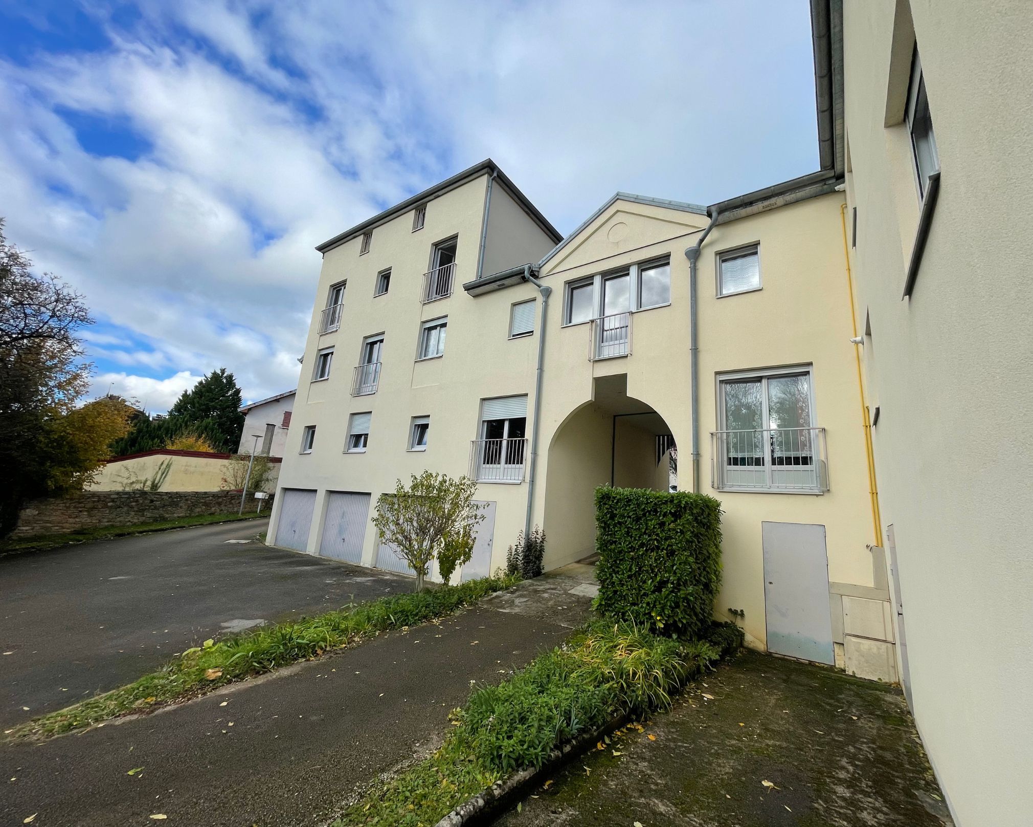 Façade d'un immeuble résidentiel beige avec plusieurs fenêtres, garages au rez-de-chaussée, une arche d'entrée, arbustes et un chemin d'accès en pavé sous un ciel partiellement nuageux.